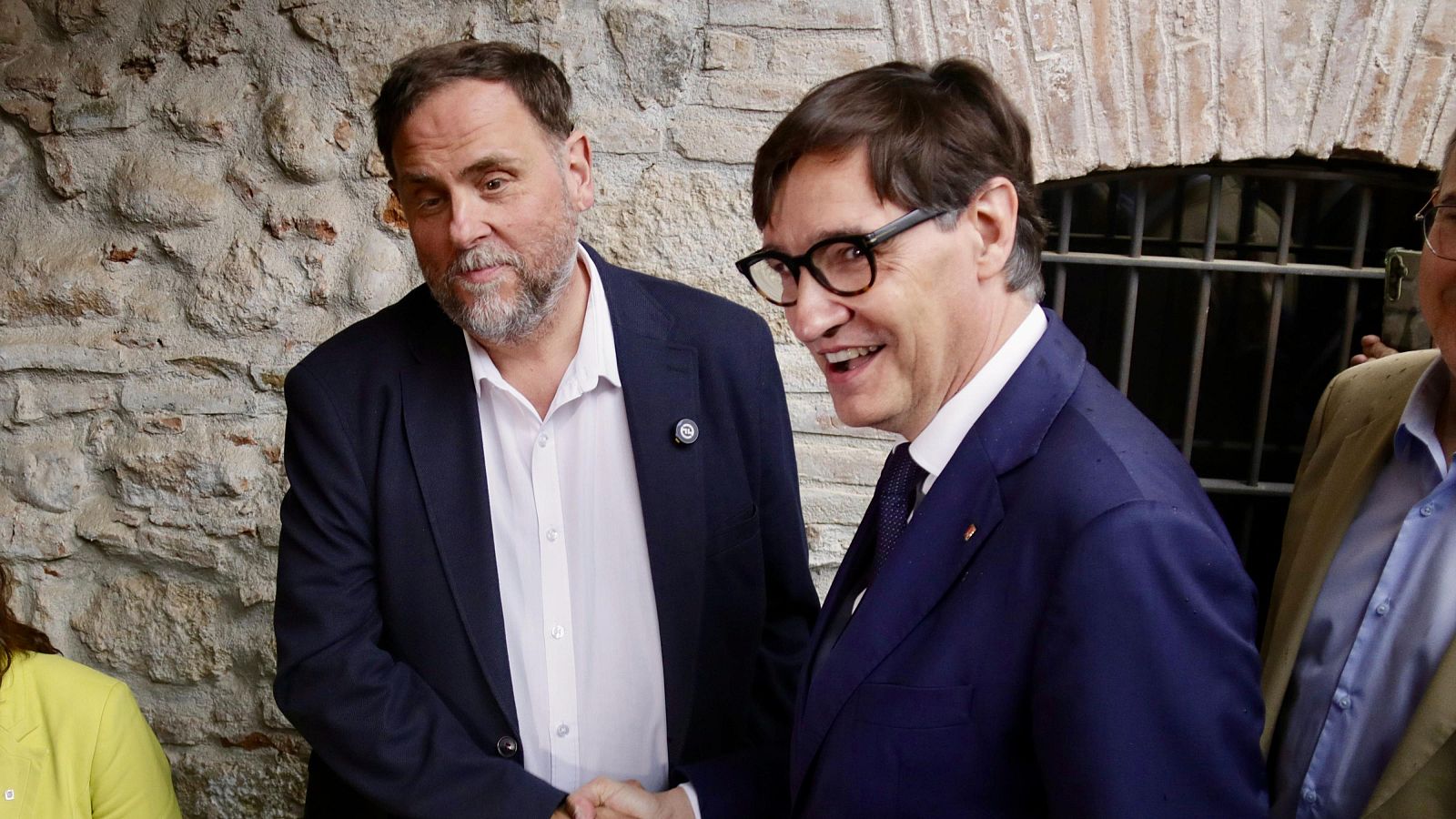 Oriol Junqueras y Salvador Illa se dan la mano en un evento político. Junqueras viste traje azul marino y barba, Illa lleva gafas y traje azul con corbata. El fondo es una pared de piedra rústica.