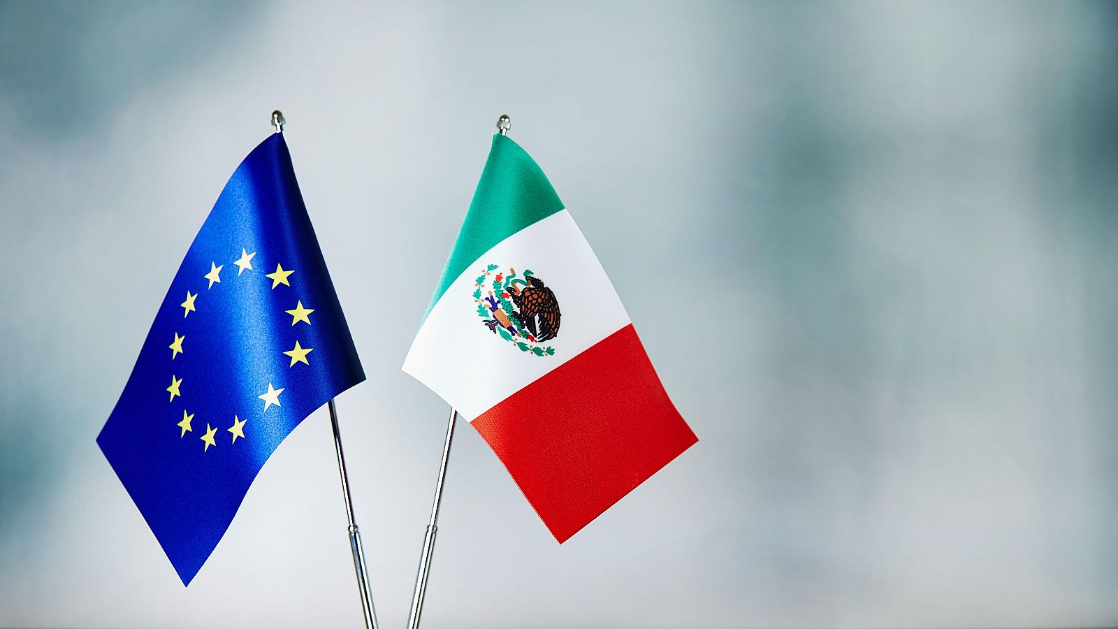 Banderas de la Unión Europea y México sobre mástiles de escritorio. La bandera de la UE tiene estrellas doradas sobre fondo azul. La de México exhibe el escudo nacional en su franja blanca central.