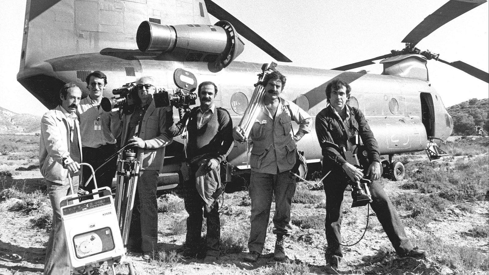 Miguel de la Quadra-Salcedo y equipo de TVE junto a un helicóptero en un terreno árido. Hombres con bigote y equipo de filmación.