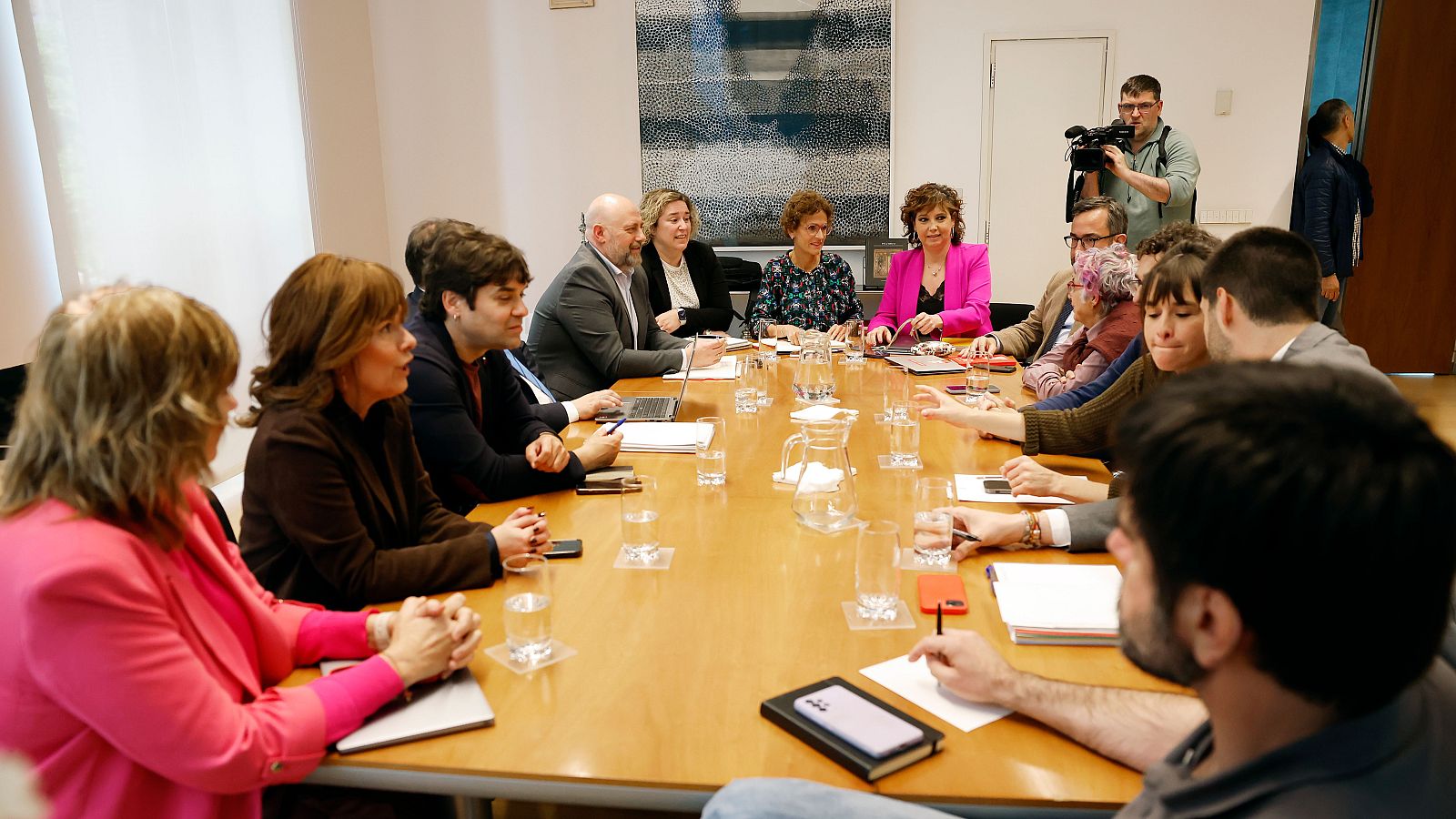 Reunión formal en sala de conferencias con María Chivite y Unai Hualde. Portátiles, documentos y vasos sobre la mesa. Camarógrafo graba el evento.