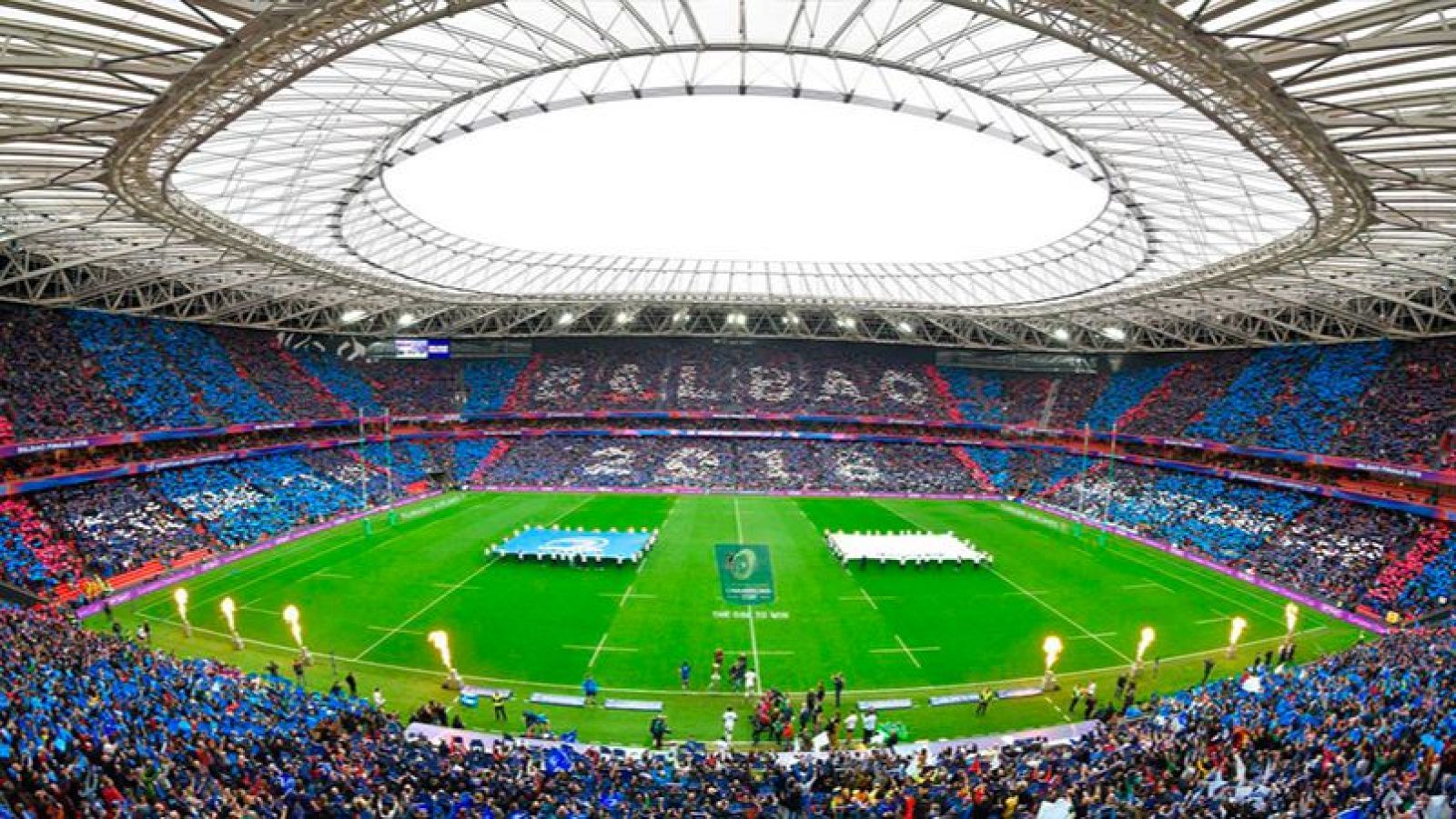 Estadio de rugby lleno en Bilbao en 2018, con gradas formando "BILBAO" y "2018", y lanzallamas activos en el campo.