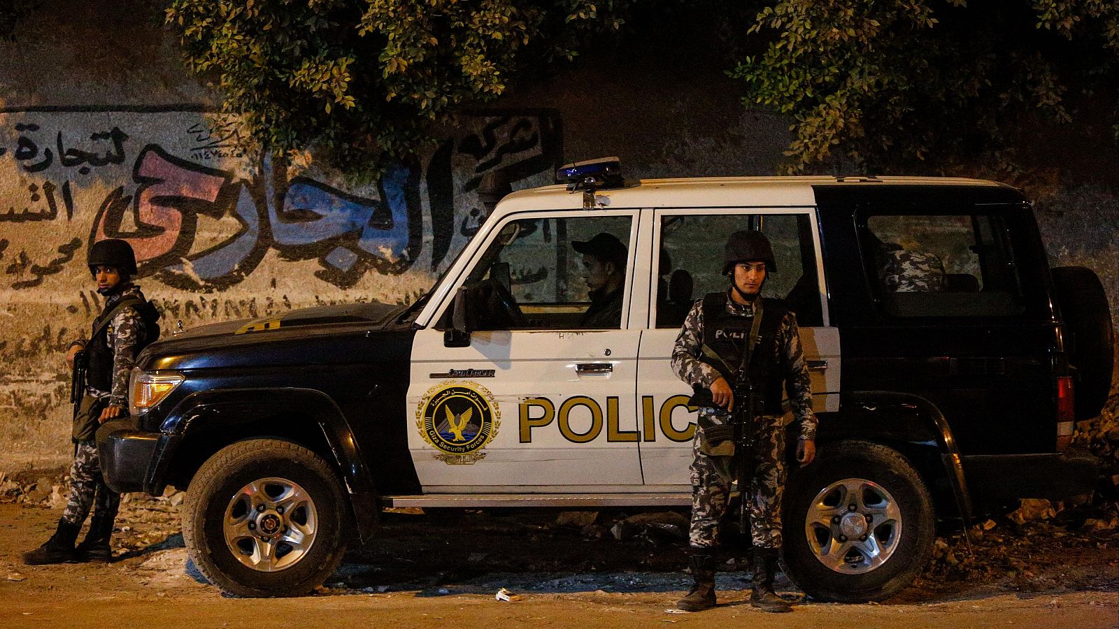 Vehículo policial "POLICE" blanco y negro, flanqueado por dos agentes armados con uniformes de camuflaje y cascos, en una calle nocturna con muro de grafitis árabe. Diciembre 2018, Giza, El Cairo, Egipto.