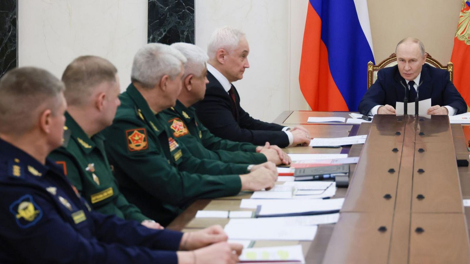 Vladímir Putin, presidente de Rusia, en una reunión formal, revisa documentos. A su lado, la bandera rusa y un estandarte estatal. Hombres con uniformes militares le acompañan.