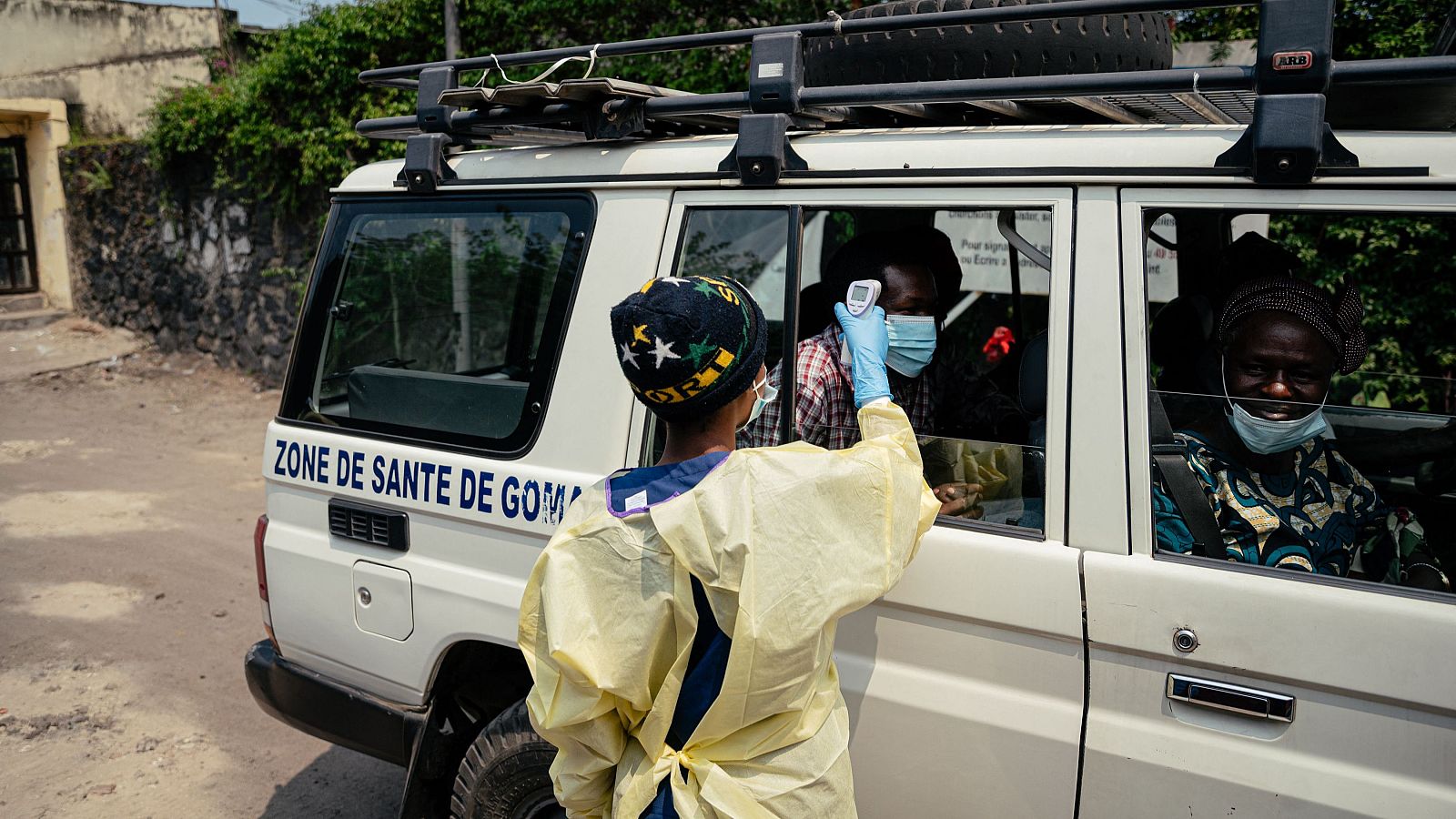 Trabajador sanitario con EPP toma temperatura a ocupantes de vehículo blanco en RDC. "ZONE DE SANTE DE GO..." se lee en el lateral. Contexto: brote de ébola.