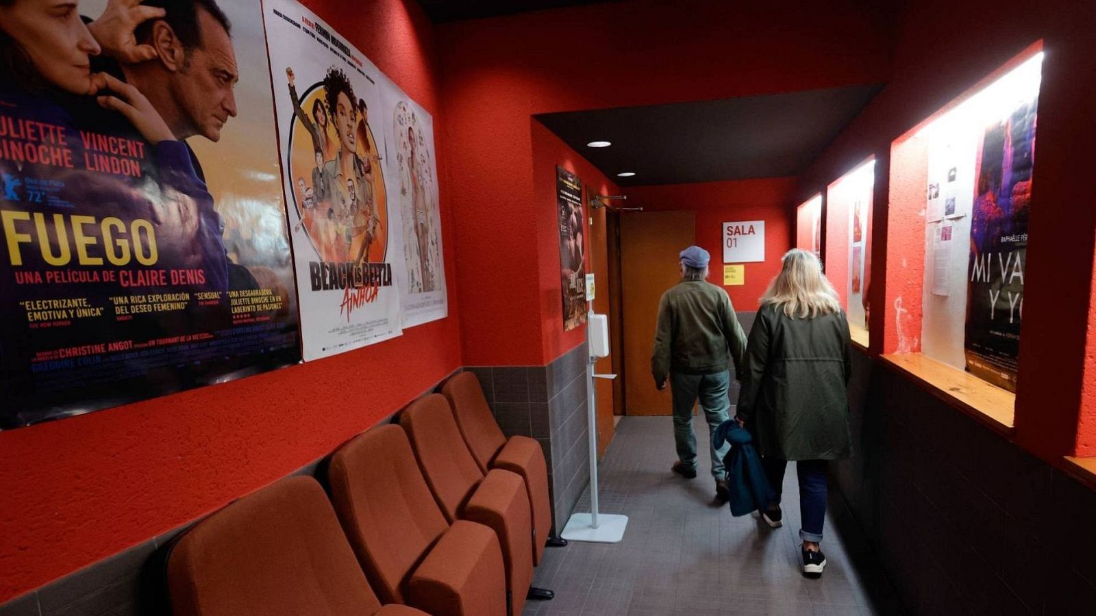 Pasillo de cine con paredes rojas, asientos naranjas y carteles de "FUEGO" y "BEAU IS AFRAID". Dos personas caminan de espaldas hacia la "SALA 01".