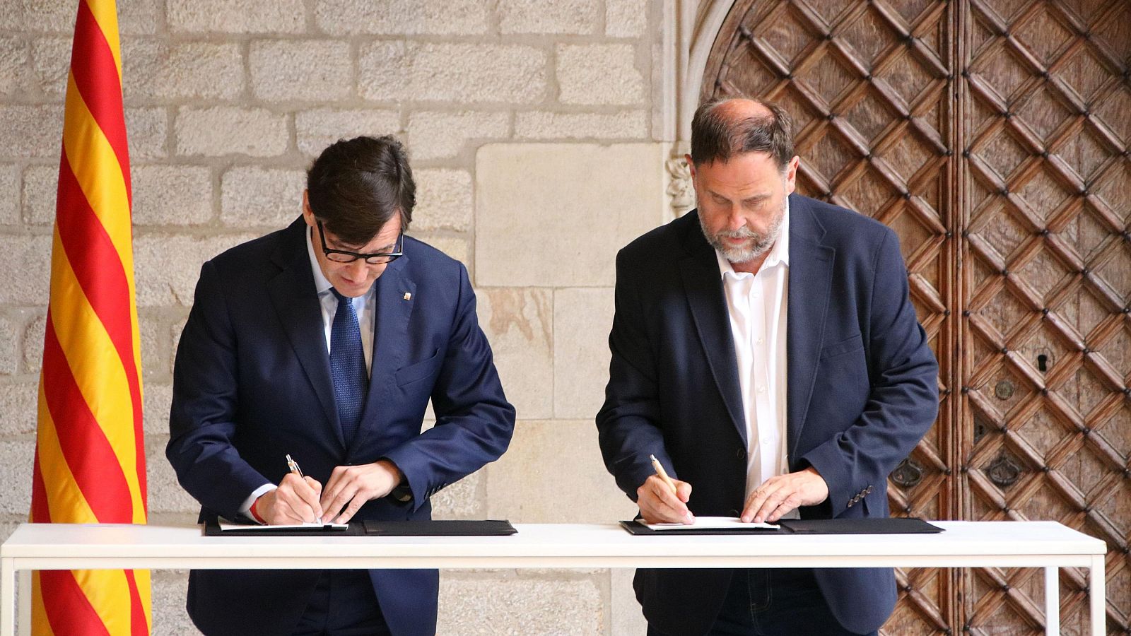 Salvador Illa y Oriol Junqueras firman documentos en un acto oficial. Illa viste traje azul marino, Junqueras americana azul. Se aprecian la Senyera y una puerta de madera tallada.