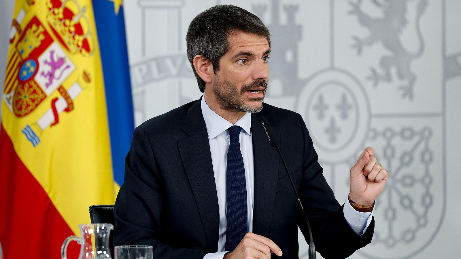 Primer plano de Ernest Urtasun, ministro de Cultura, en rueda de prensa. Viste traje oscuro, camisa blanca y corbata azul. Se ve la bandera de España y un emblema oficial. Sostiene un micrófono.