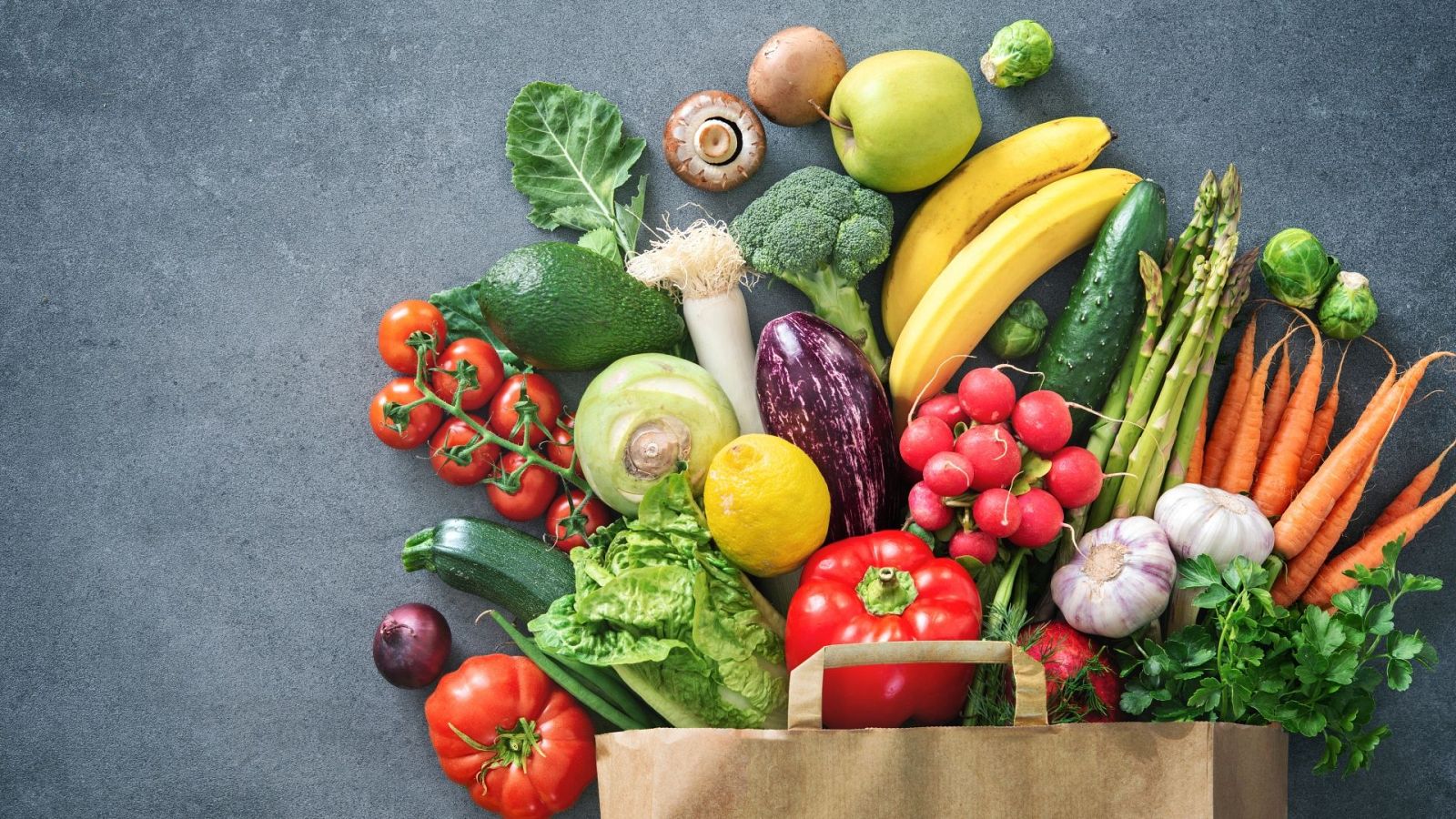 Gran variedad de frutas y verduras frescas, como tomates, aguacates, limones, plátanos, cebollas, pimientos, lechugas, brócoli, zanahorias y ajos, desbordándose de una bolsa de papel marrón sobre fondo texturizado gris.
