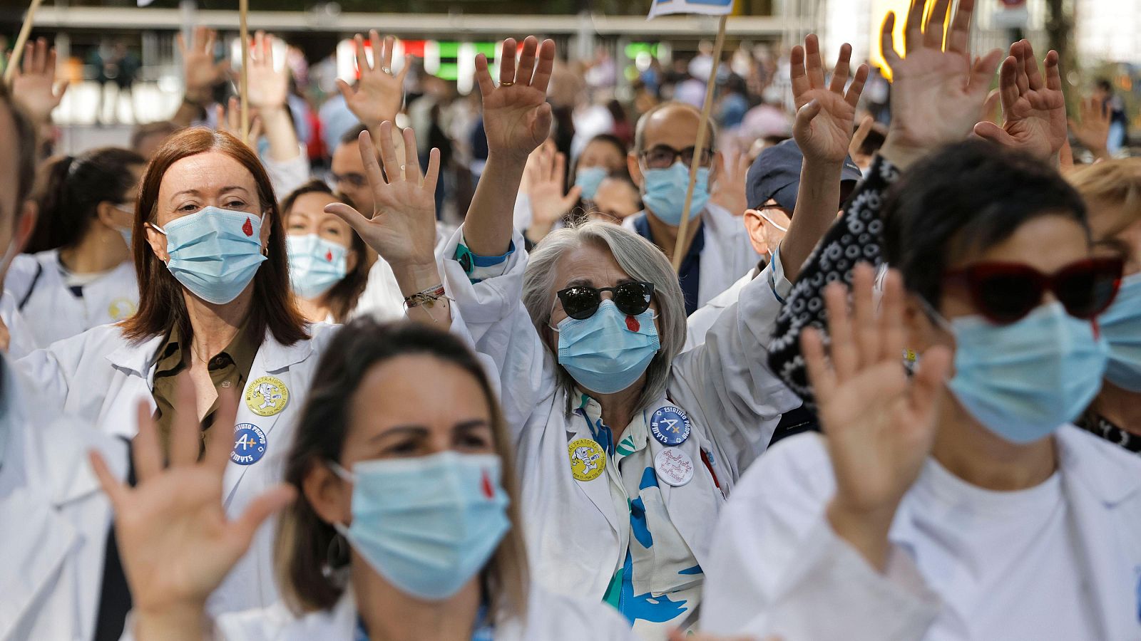 Médicos en huelga con batas blancas y mascarillas levantan las manos en protesta. Se aprecian chapas y símbolos de gotas de sangre en algunas mascarillas.