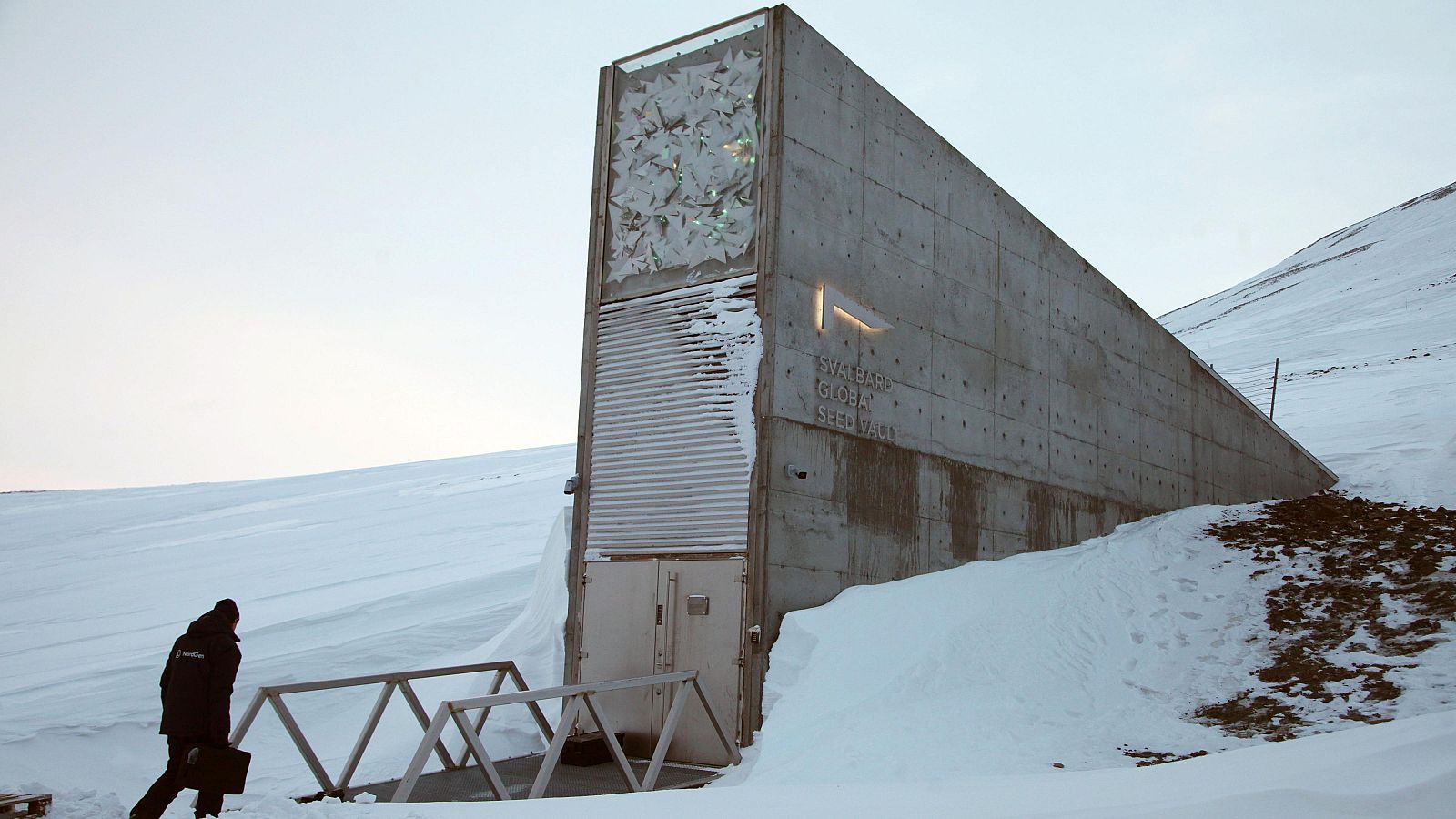 Vista exterior de la Bóveda Global de Semillas de Svalbard, con un visitante dirigiéndose a su entrada bajo un paisaje nevado. El letrero "SVALBARD GLOBAL SEED VAULT" es visible en la fachada.
