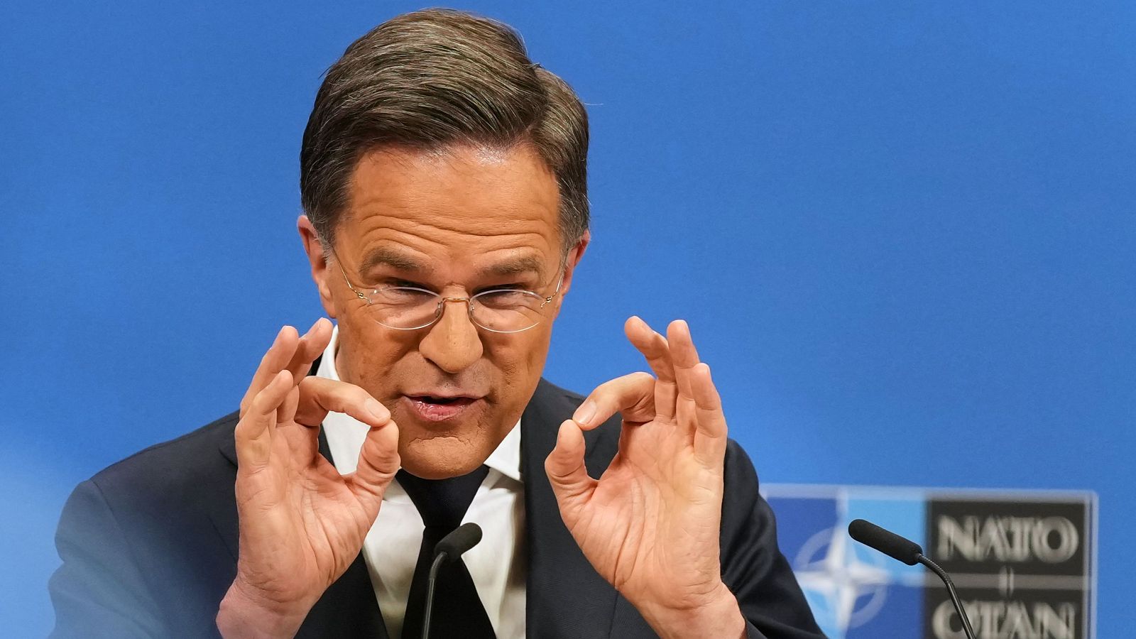 Mark Rutte, Primer Ministro de los Países Bajos, hace el gesto de "OK" frente al logo de la OTAN. Lleva traje oscuro, camisa blanca y gafas.