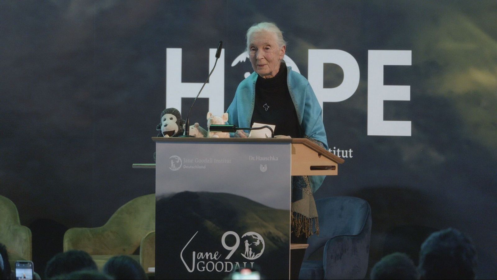 Jane Goodall, vestida con un chal azul turquesa, habla en un evento. Detrás, "HOPE" en un fondo nublado. El atril muestra "Jane GOODALL 90" y logos de "Jane Goodall Institut Deutschland" y "Dr. Hauschka".
