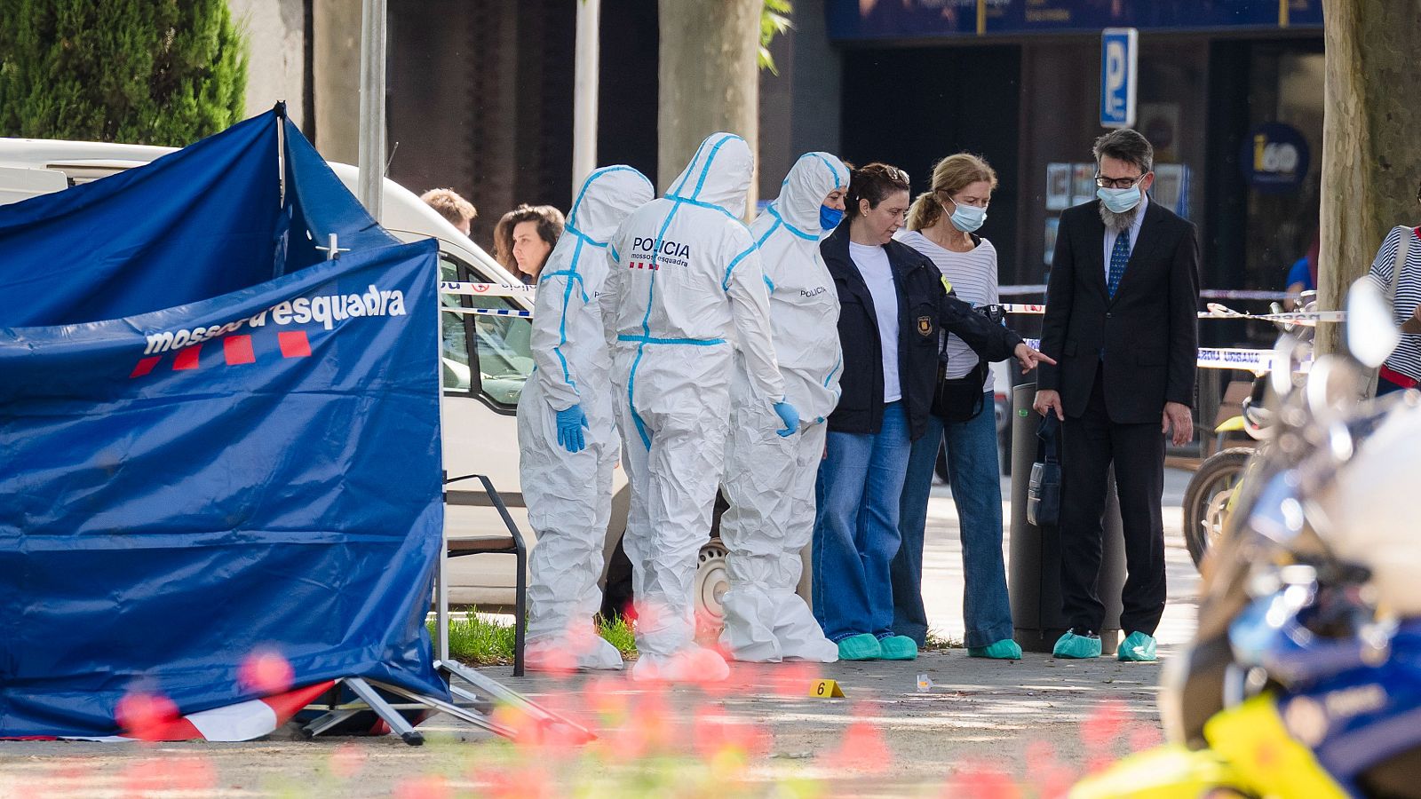 Investigación policial de los Mossos d'Esquadra en una plaza urbana. Agentes forenses y otros participantes examinan la evidencia en el suelo, marcada con '6 A' y delimitada por cinta policial.