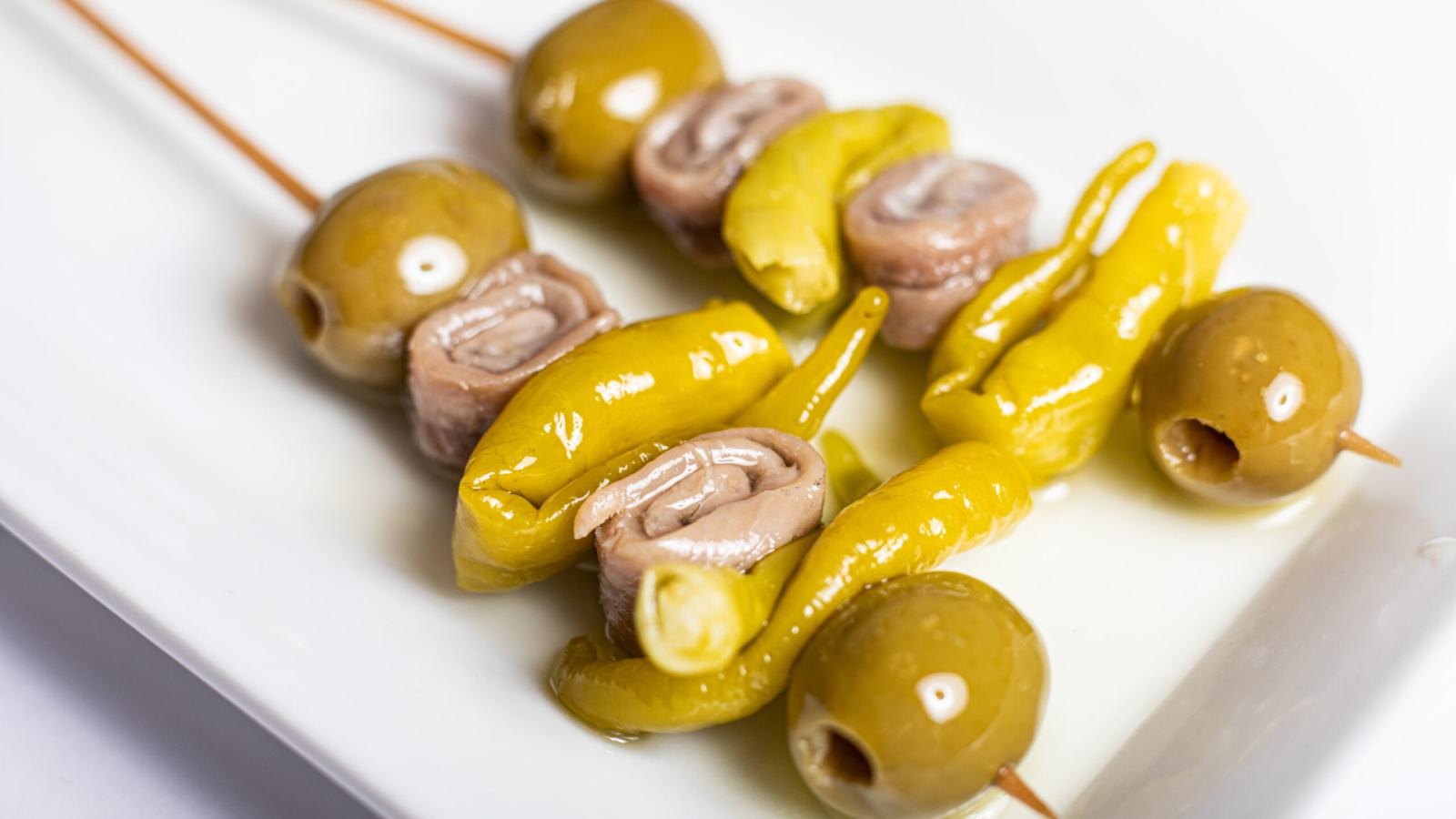 Primer plano de gildas sobre plato blanco: aceituna verde, anchoa enrollada y guindilla en palillo. Detalle de ingredientes y líquido en la base.