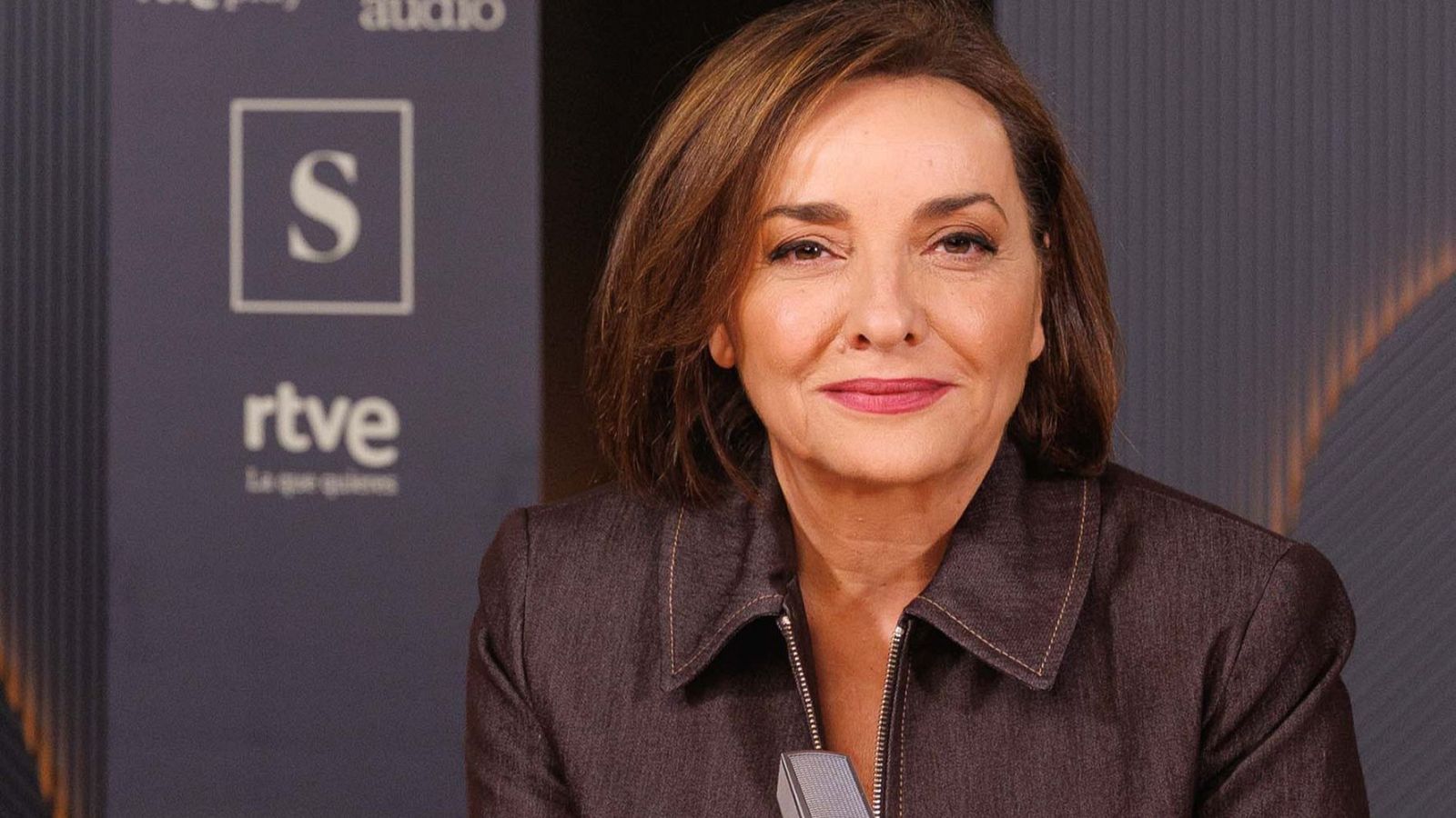 Retrato de Pepa Bueno, periodista española, con chaqueta oscura y fondo con logotipos de RTVE y elementos de audio.