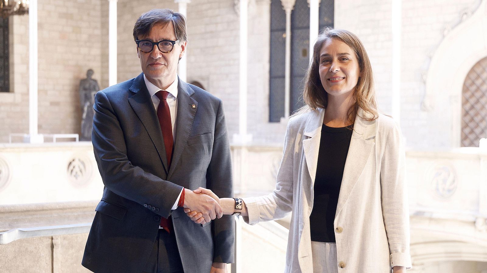 Salvador Illa i Jéssica Albiach, líders polítics, formalitzen un acord amb una encaixada de mans davant d'una arquitectura de pedra.