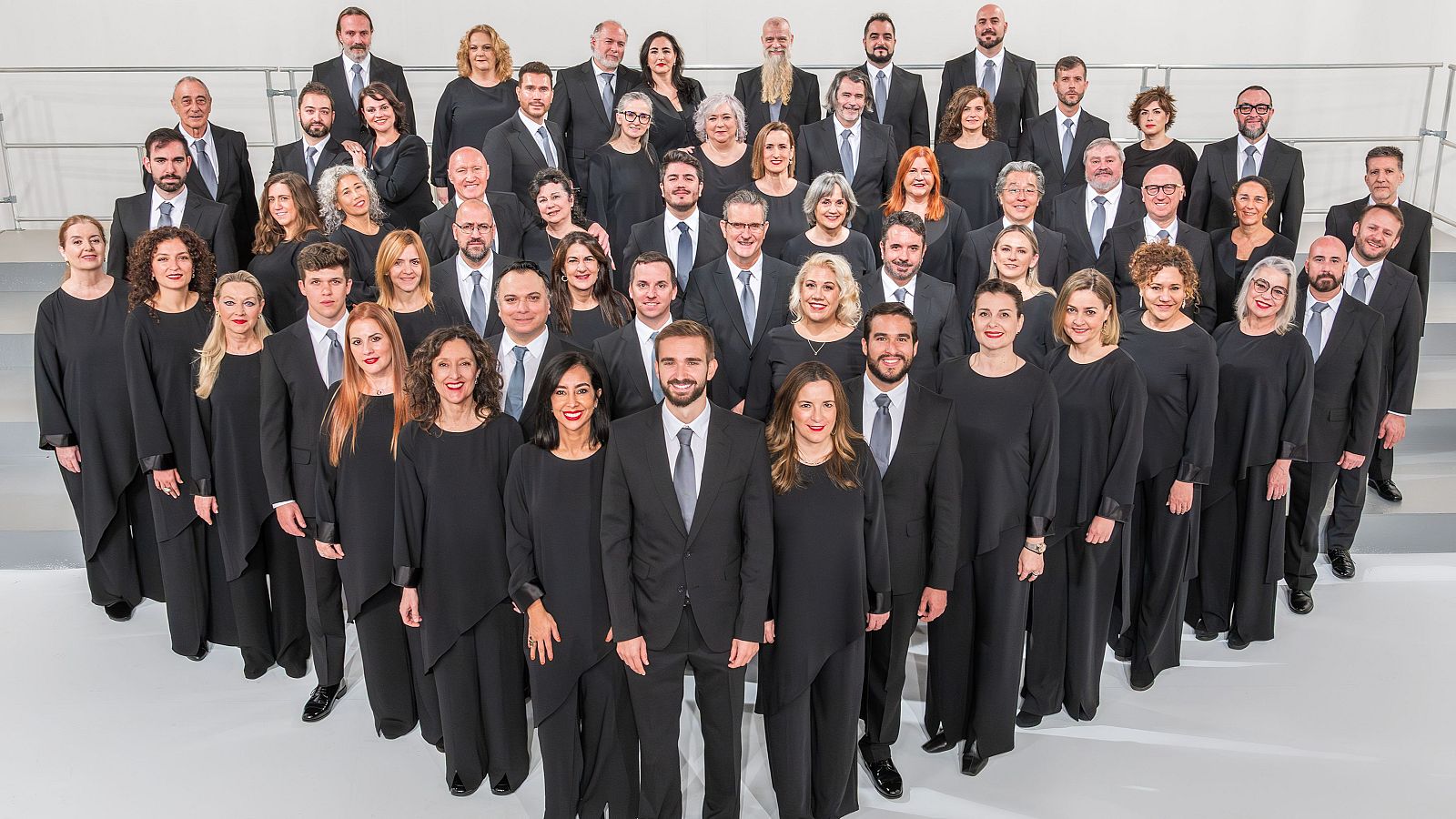 El Coro de RTVE, un grupo de 55-60 adultos con vestimenta formal y uniforme (trajes oscuros para hombres, túnicas negras para mujeres), posa en filas escalonadas sobre un fondo liso y claro.