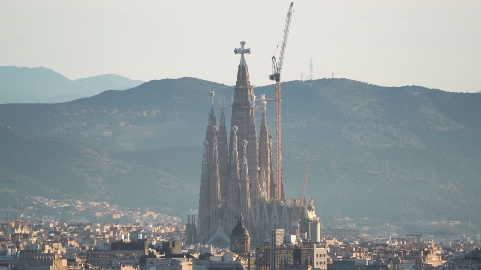 Vista panoràmica de la Sagrada Família a Barcelona, amb les seves torres i una grua de construcció. Al voltant, la ciutat i muntanyes al fons.