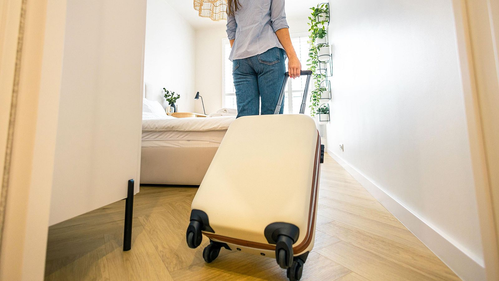 Mujer de espaldas con camisa azul y vaqueros, arrastra maleta beige por habitación con suelo de madera, cama y jardín vertical.
