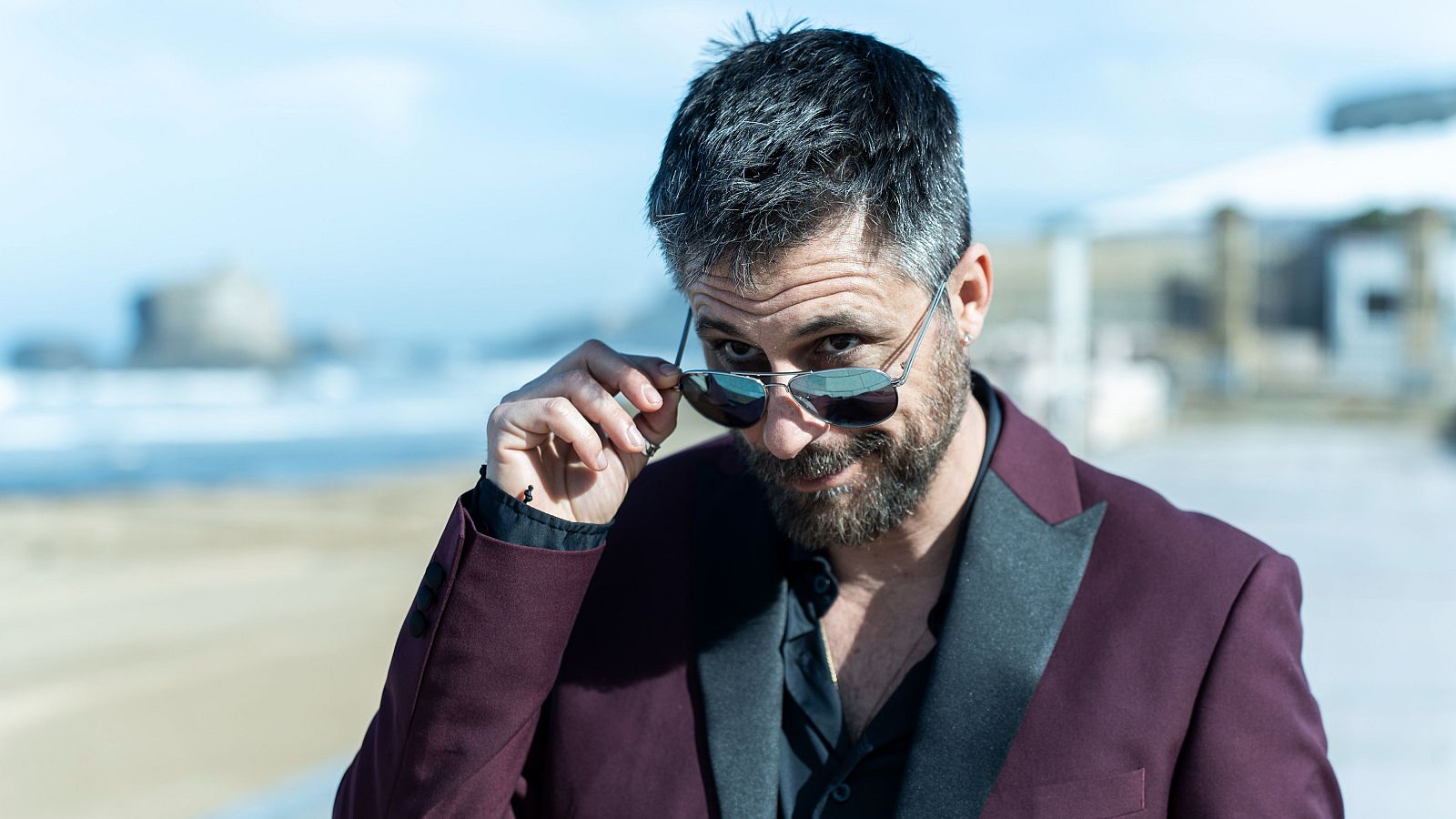 Primer plano de Hugo Silva con gafas de aviador, chaqueta burdeos y camisa oscura, con un paisaje costero de fondo.