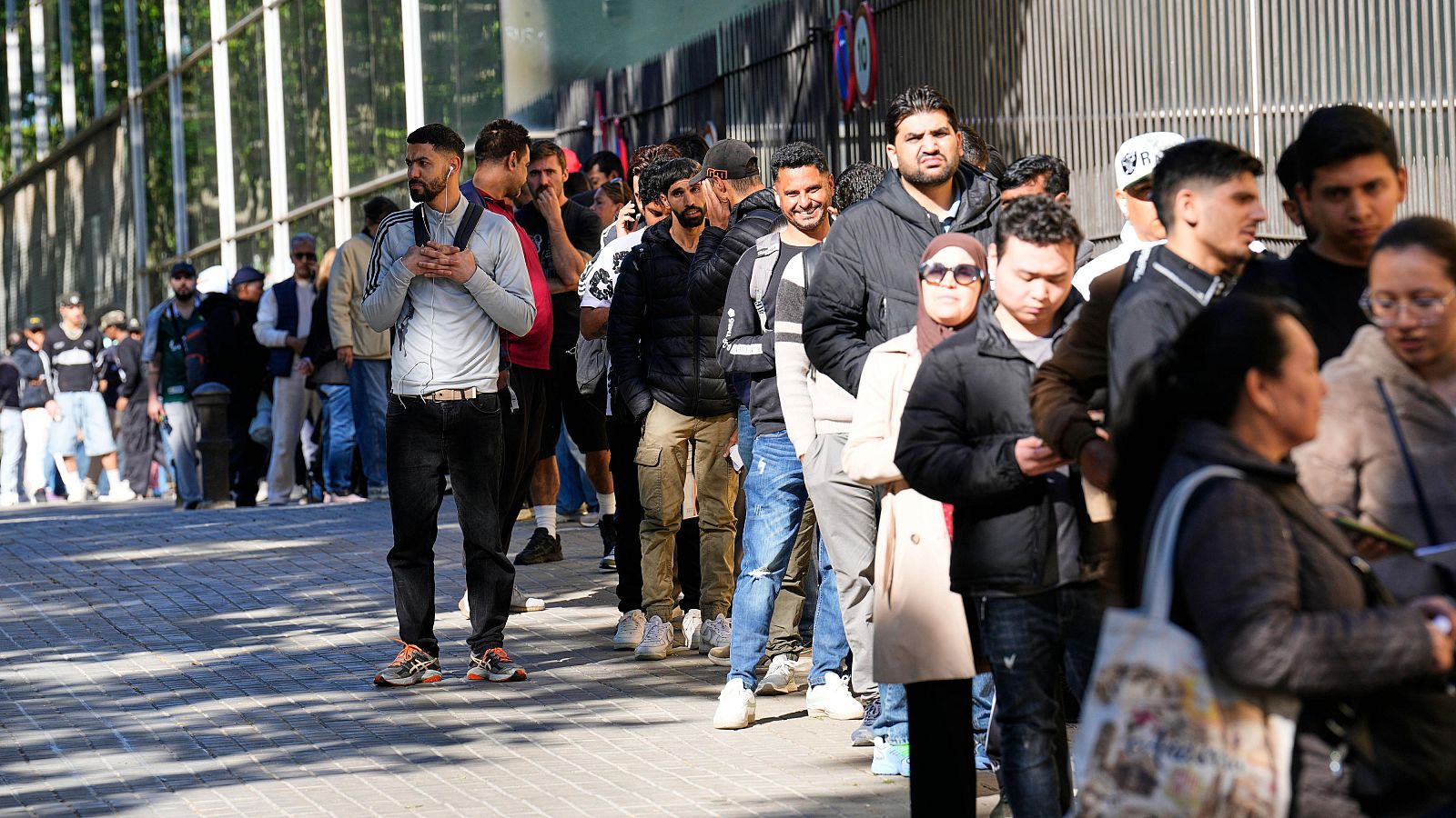 Fila de personas migrantes en Barcelona esperando al aire libre en un día soleado para realizar un trámite de regularización.