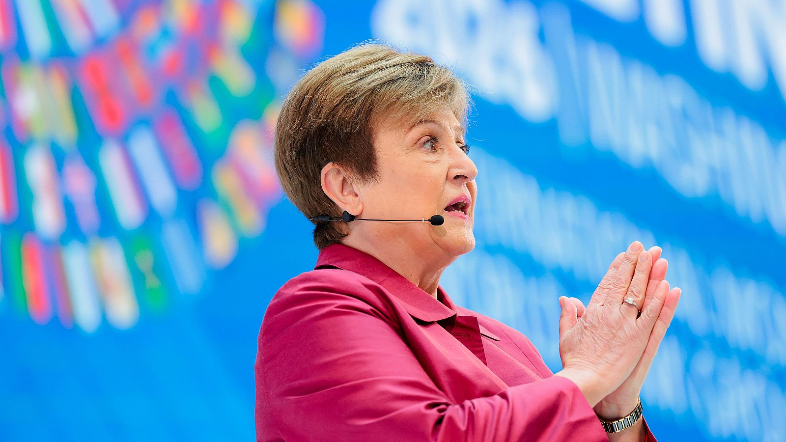 Kristalina Georgieva, directora gerente del FMI, habla con un micrófono de diadema y chaqueta fucsia, con fondo azul desenfocado y elementos gráficos.