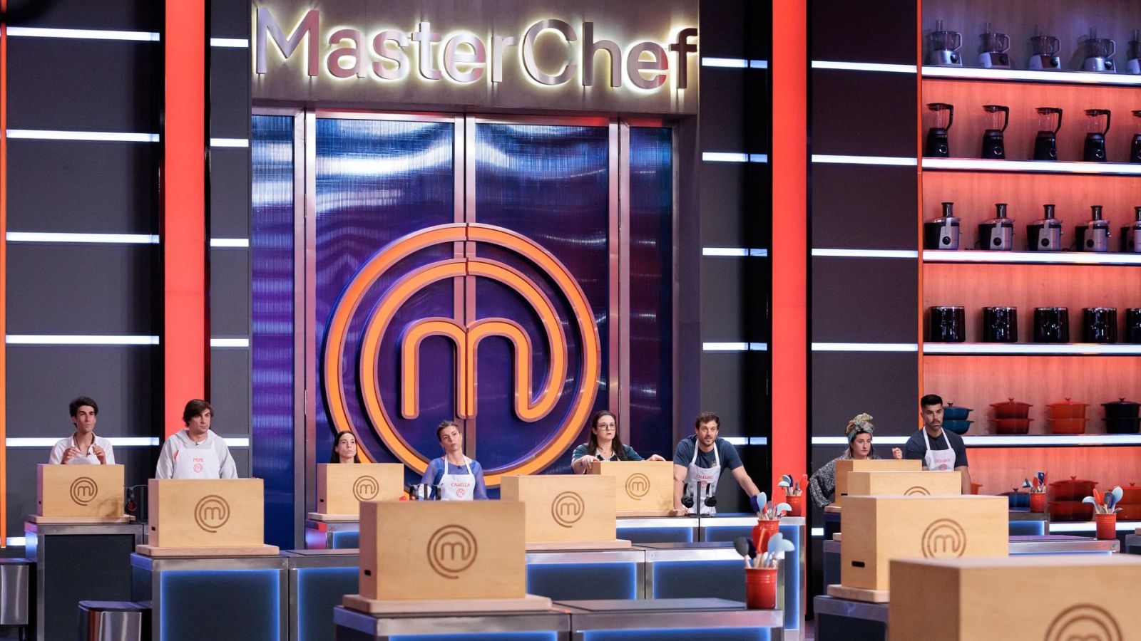 Plató de MasterChef con siete aspirantes en sus estaciones de cocina, cada uno con una caja de ingredientes. El logo del programa es visible en el fondo.