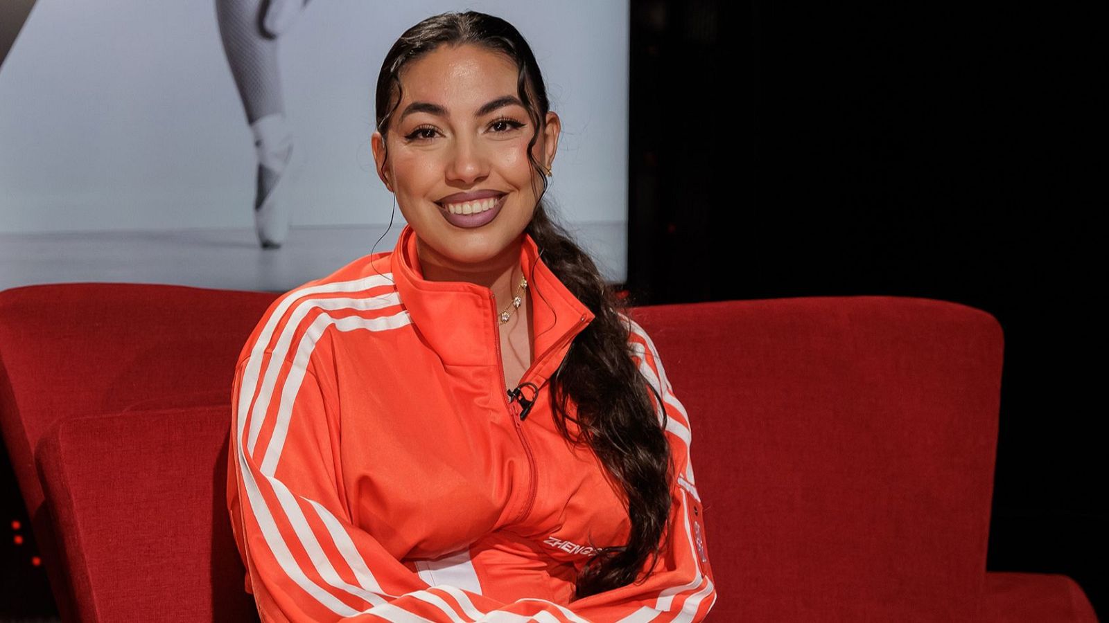 María José Llergo sonríe en un sofá rojo, vistiendo una chaqueta deportiva naranja con franjas blancas. El fondo es mixto, con una proyección de danza y oscuridad.