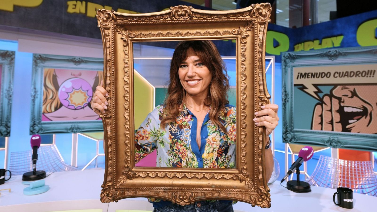 Nuria Marín en "¡Menudo Cuadro!" con un marco dorado, rodeada de arte pop y micrófonos de radio. "EN PLAY" se ve al fondo.