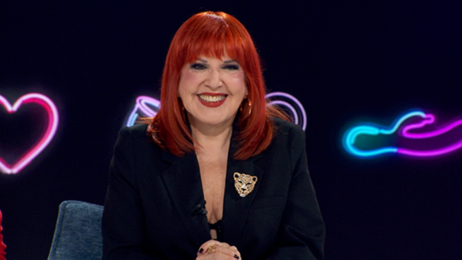 Loles León, con pelo rojo y blazer negro con broche de leopardo, sonríe rodeada de luces de neón y un corazón rosa.