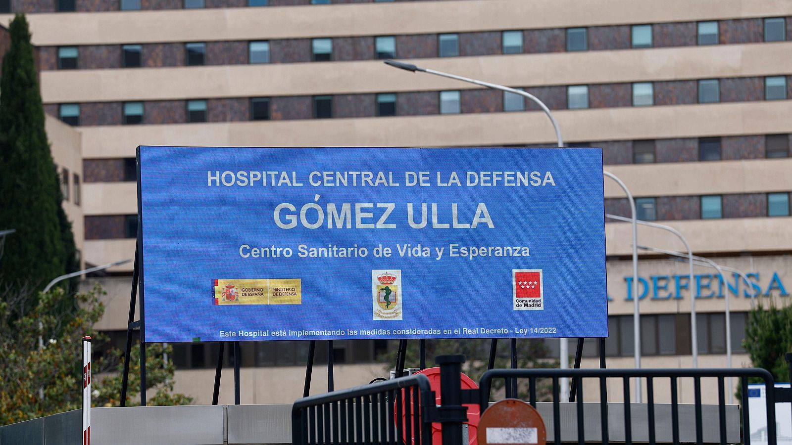 Cartel azul con "HOSPITAL CENTRAL DE LA DEFENSA GÓMEZ ULLA" y logos institucionales frente al edificio del Hospital Gómez Ulla. Se ve un árbol y una valla en primer plano.