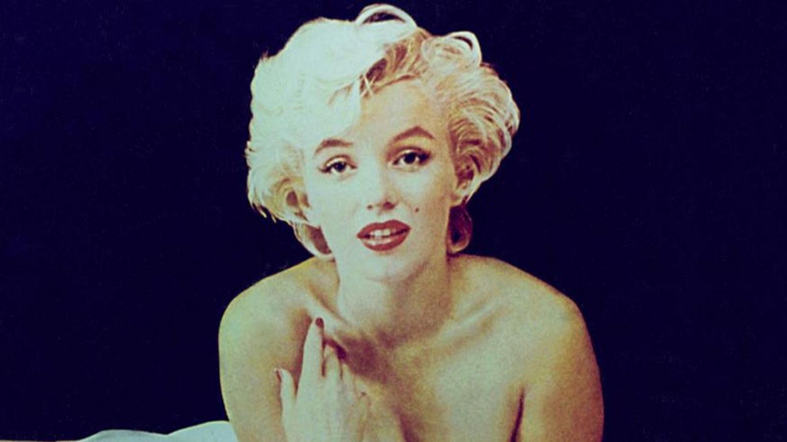 Retrato de Marilyn Monroe con cabello rubio platino, labios rojos y lunar, apoyando su mano en el hombro sobre fondo oscuro.
