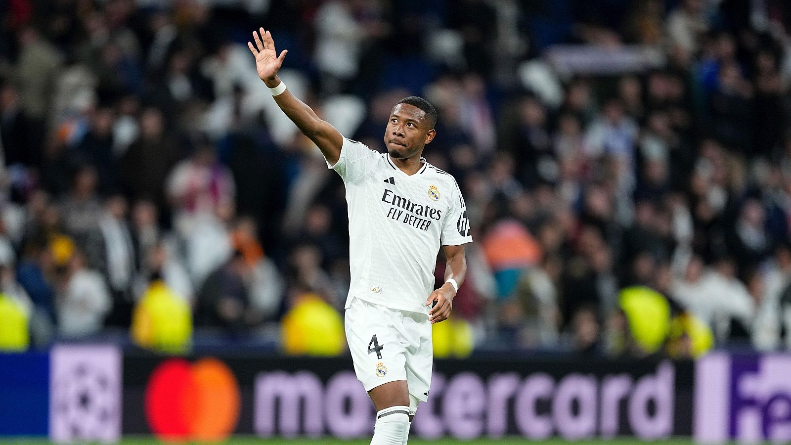 David Alaba del Real Madrid saluda con la mano levantada, luciendo la camiseta blanca con el número 4 y logos de Adidas, Emirates y el escudo del club. El estadio lleno y patrocinadores como Mastercard y la Champions League completan la escena.