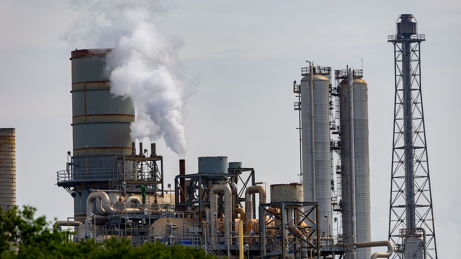 Complejo industrial con chimenea humeante, torres metálicas y red de tuberías bajo un cielo claro. Se observa vegetación en primer plano y una torre de celosía a la derecha.
