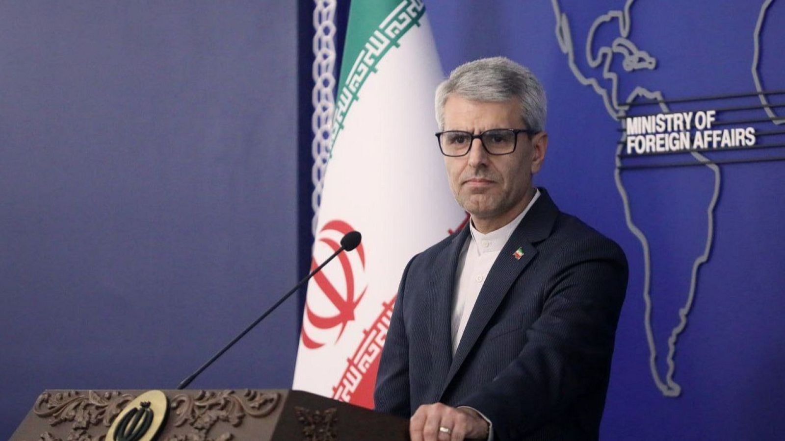 Hombre con gafas y traje oscuro, frente a un micrófono y atril con emblema iraní. Detrás, bandera de Irán y mapa del mundo con "MINISTRY OF FOREIGN AFFAIRS".