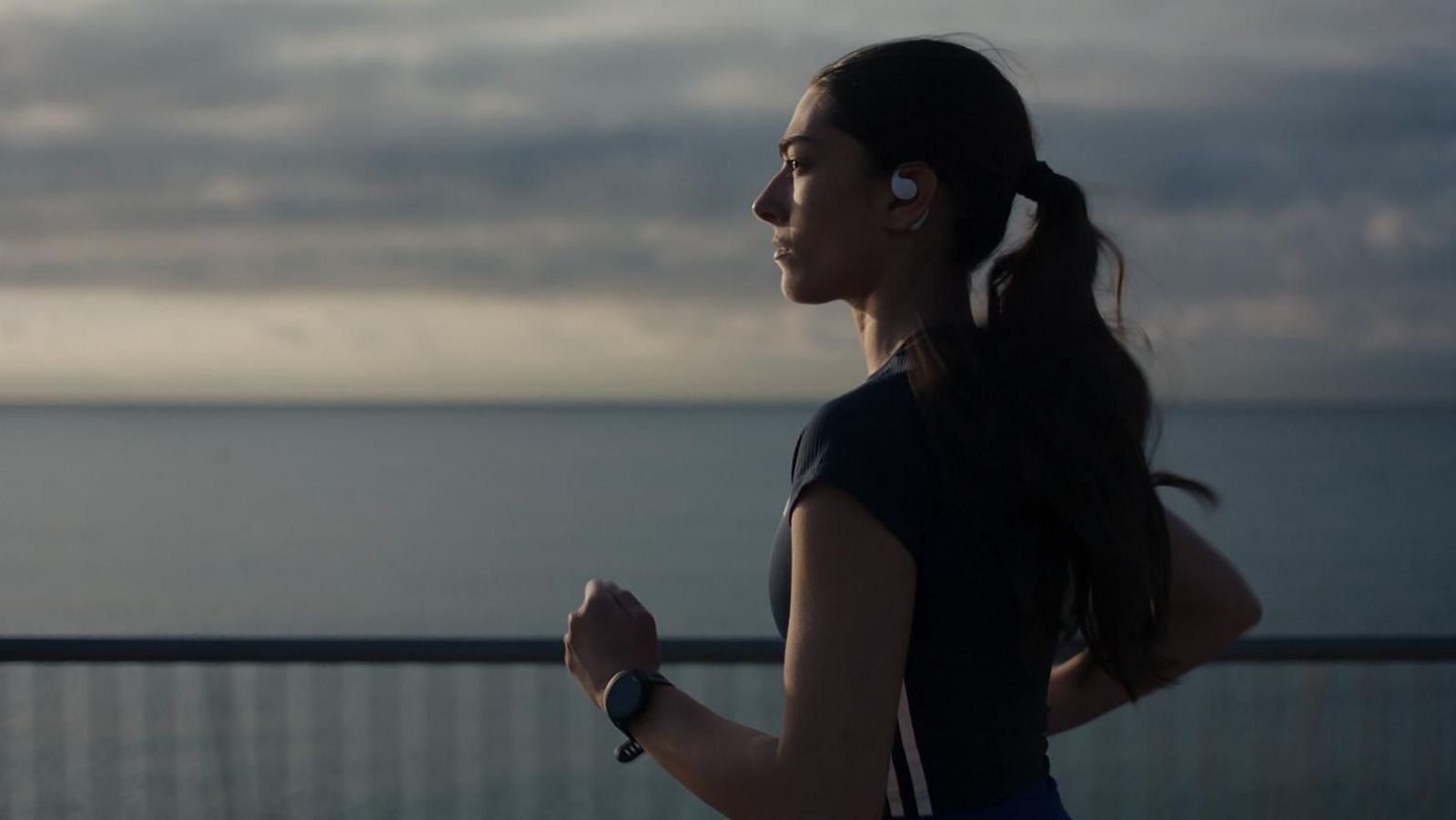Mujer joven corriendo al aire libre, de perfil, con ropa deportiva, auricular y reloj inteligente, en un entorno costero con mar y cielo nublado.