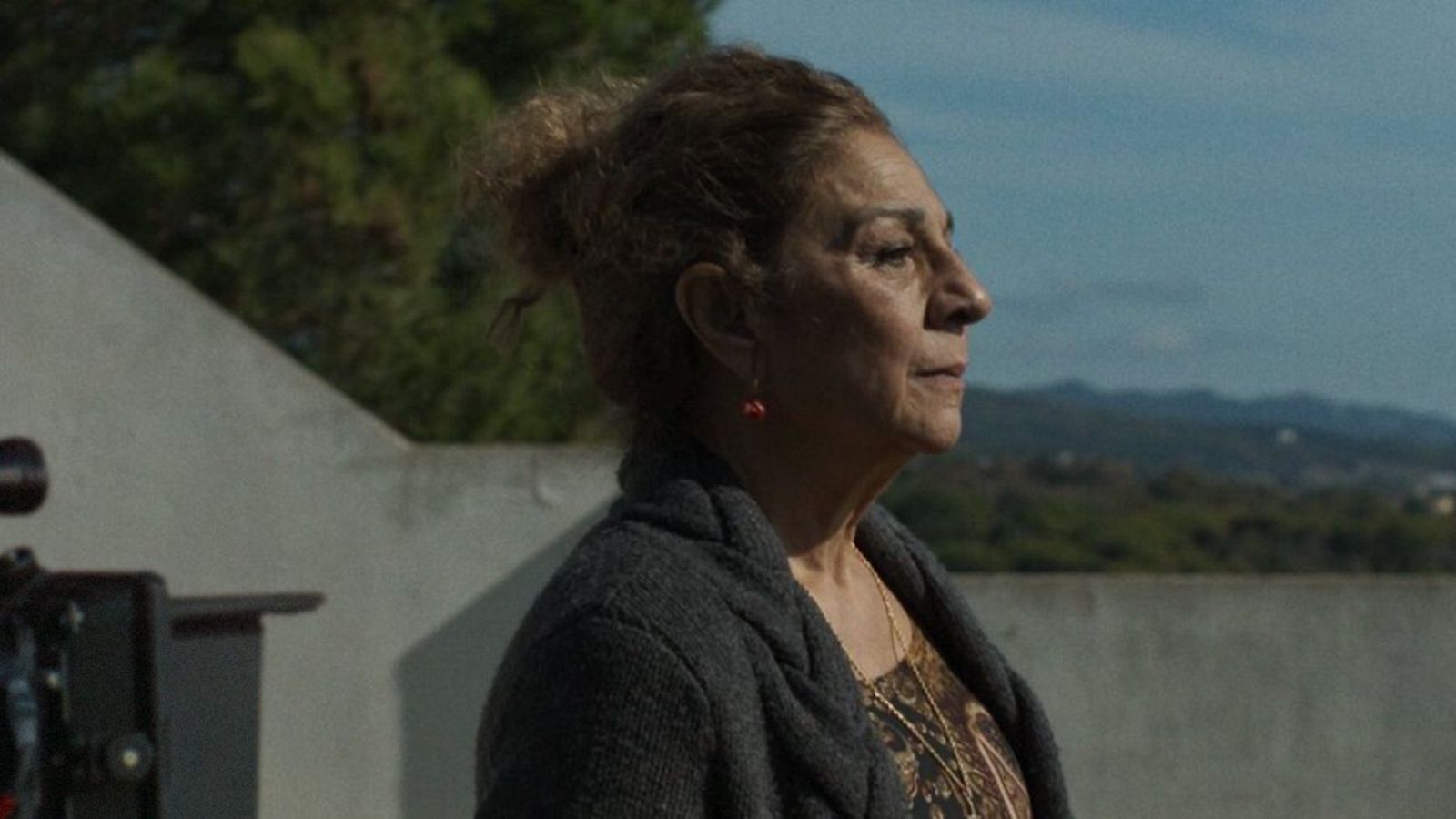 Lolita Flores de perfil, con cabello recogido, cárdigan gris y blusa estampada. Fondo de colinas verdes y cielo azul. Se intuye equipo de cámara.