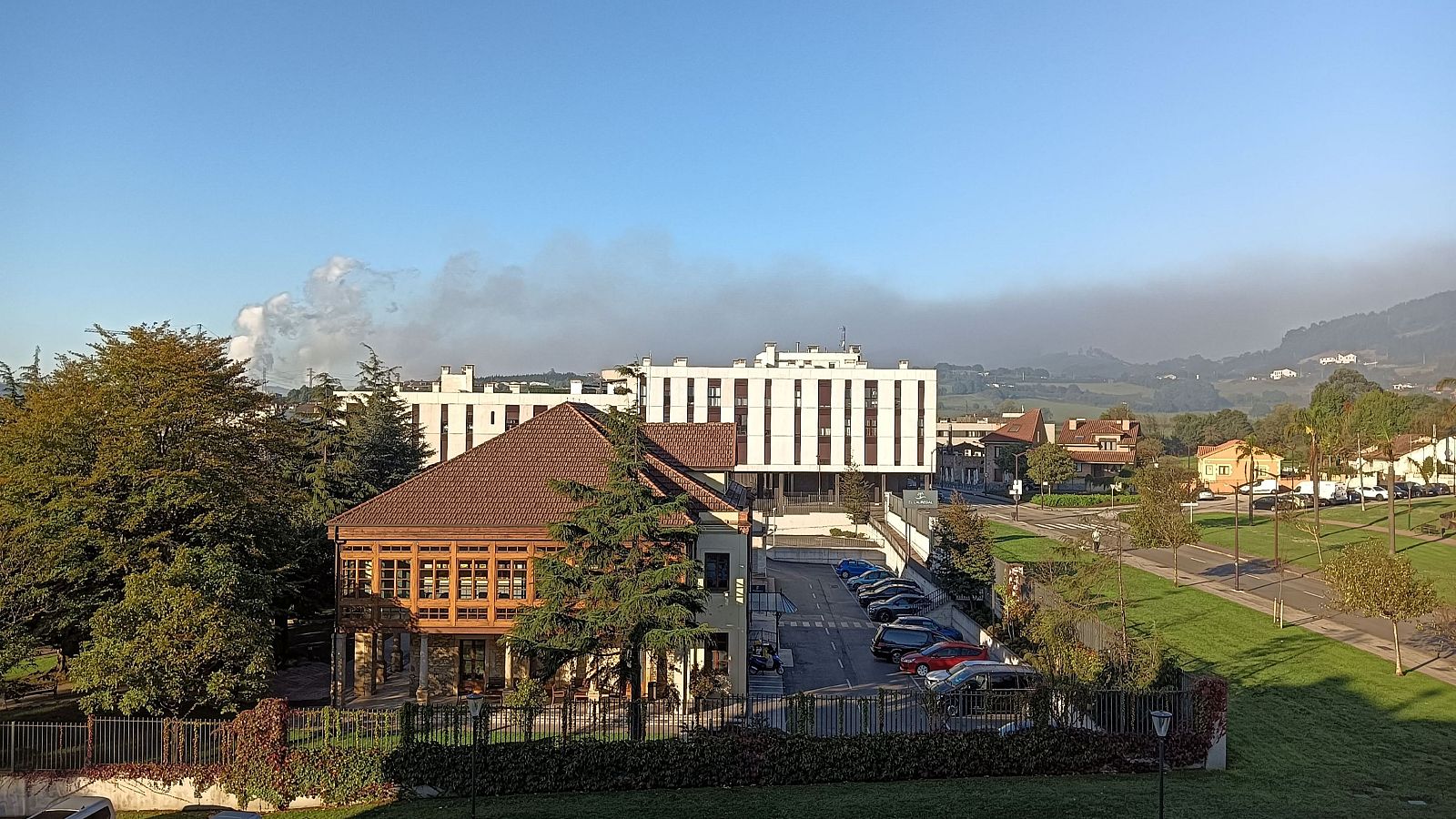 Vista de Lauredal, Gijón, Asturias, con un penacho de humo y una boina de contaminación grisácea cubriendo el horizonte. Se observan edificios, vegetación y coches.