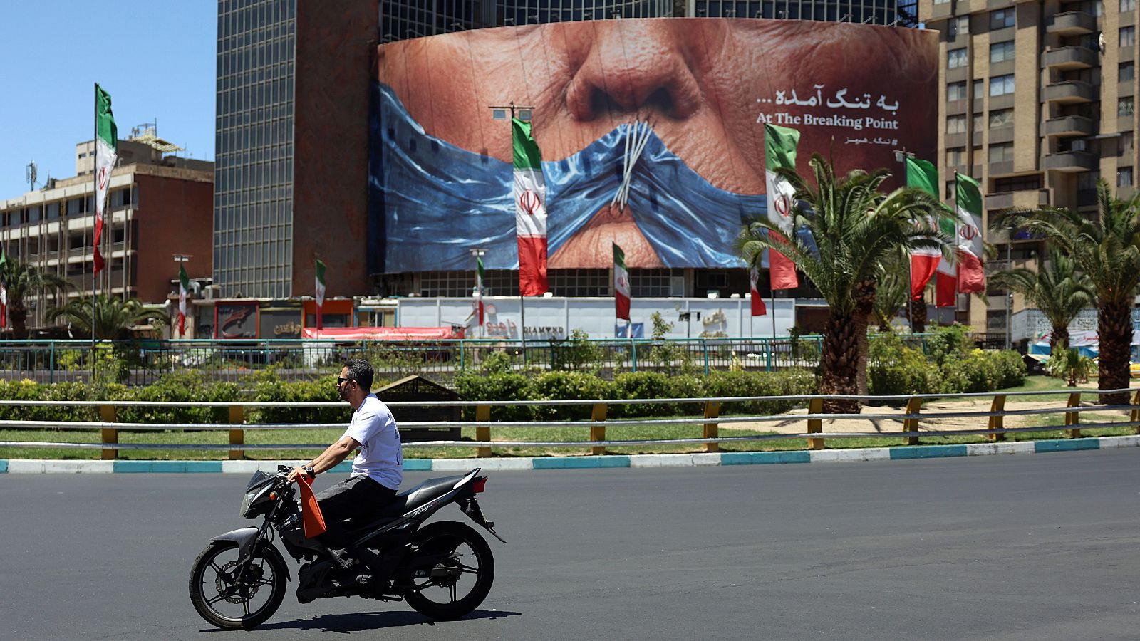 Motorista en Teherán, Irán, pasa frente a un cartel de Donald Trump con la boca tapada y el texto "At The Breaking Point".