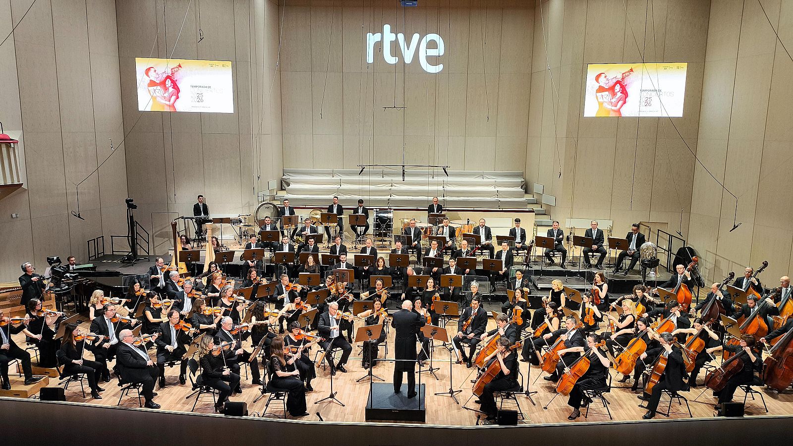 Orquesta y Coro RTVE en concierto, con director al frente, músicos y coristas en formación escalonada. Logotipo de RTVE y pantallas con arte abstracto al fondo.