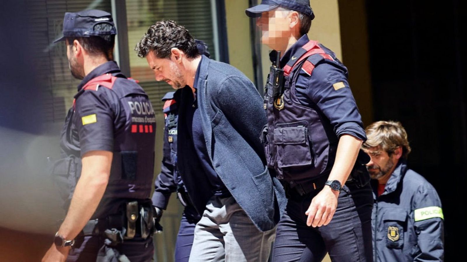 Jonathan Andic, hijo del fundador de Mango, es escoltado por agentes de los Mossos d'Esquadra. Viste traje oscuro y tiene barba y bigote. La escena sugiere un contexto legal.