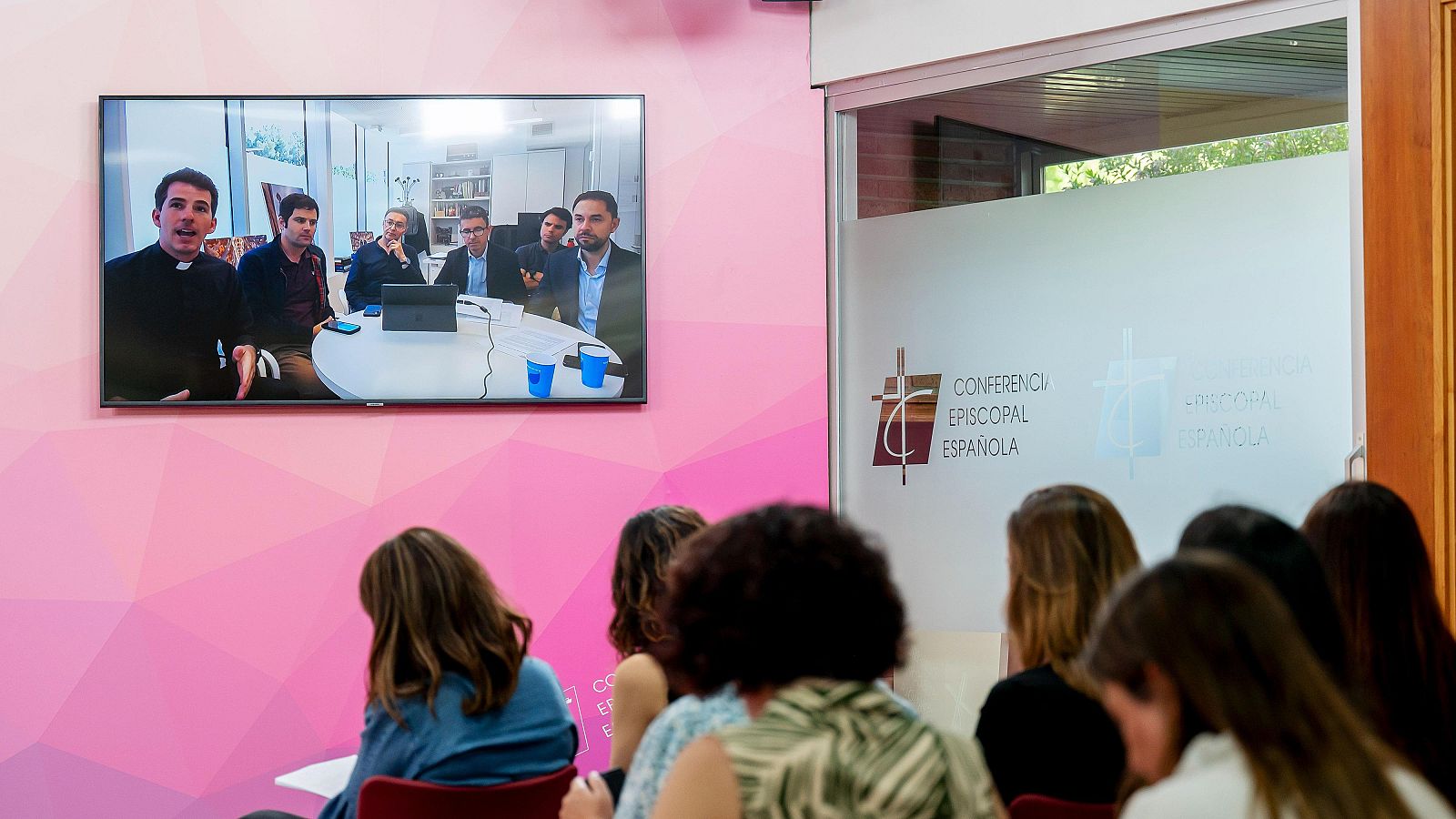 Grupo de periodistas sentados en la sala de prensa de la Conferencia Episcopal Española en Madrid mirando una gran pantalla de televisión donde aparecen los responsables de la organización de la visita del papa a Barcelona
