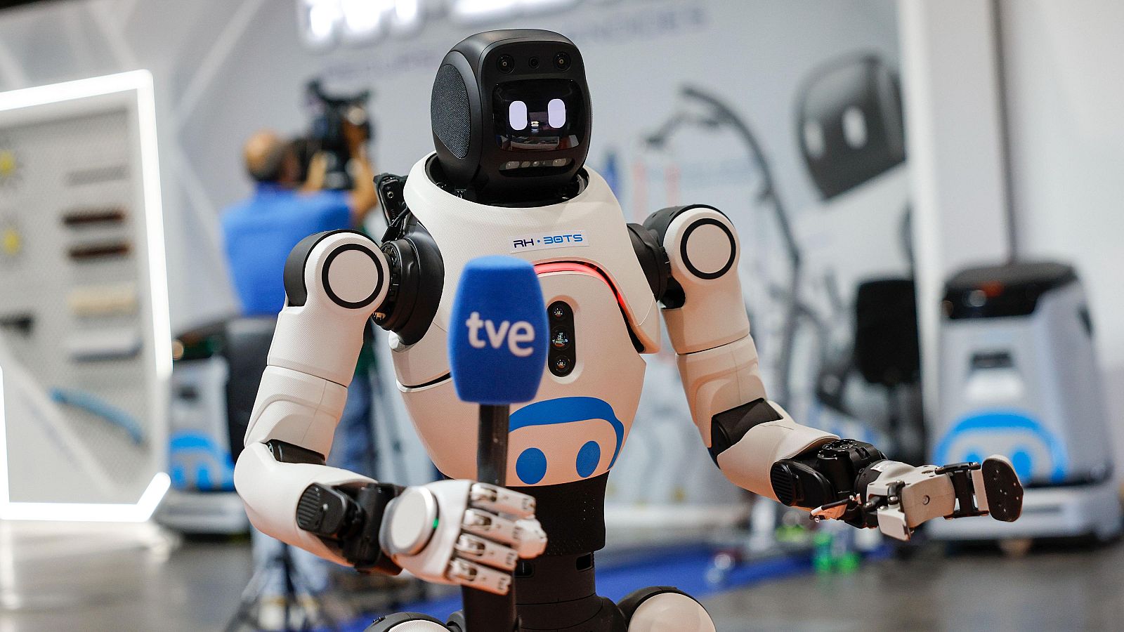 Robot humanoide "PH-BOTS" con micrófono "tve" en feria tecnológica. Fondo con cámara y otros robots.