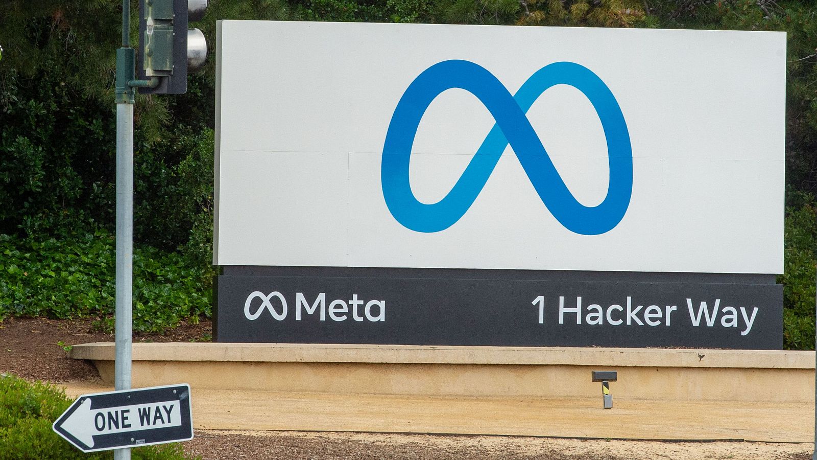 Cartel corporativo de Meta con logo de infinito azul, "Meta" y "1 Hacker Way" en letras blancas. Señal "ONE WAY" y vegetación verde al exterior.