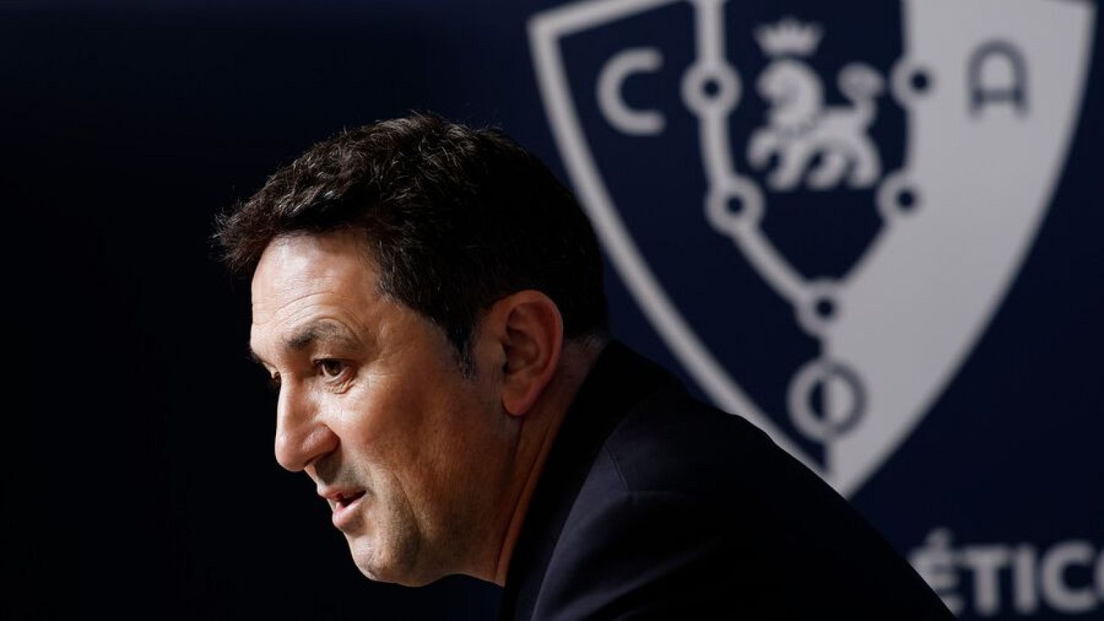 Braulio Vázquez de perfil, con expresión seria, frente al escudo del Club Atlético Osasuna que incluye las letras 'C', 'A' y un león rampante.