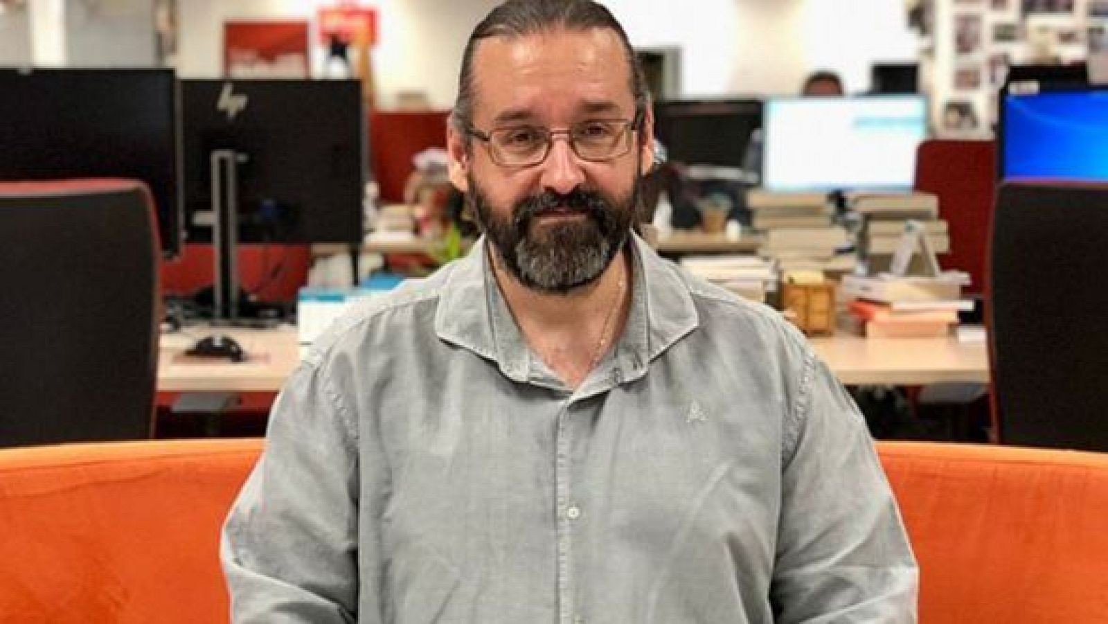 Retrato de Ramón Paso, dramaturgo, con gafas oscuras, barba y camisa grisácea, sentado en un sofá naranja. Fondo de oficina con escritorios y monitores.