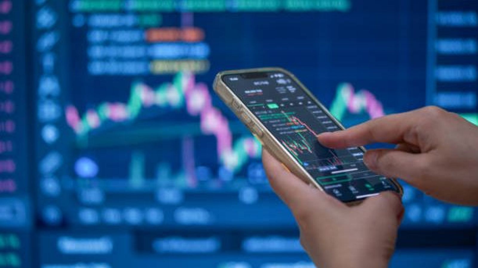 Mano con smartphone dorado muestra gráficos financieros. Fondo con pantalla grande de datos de mercado.