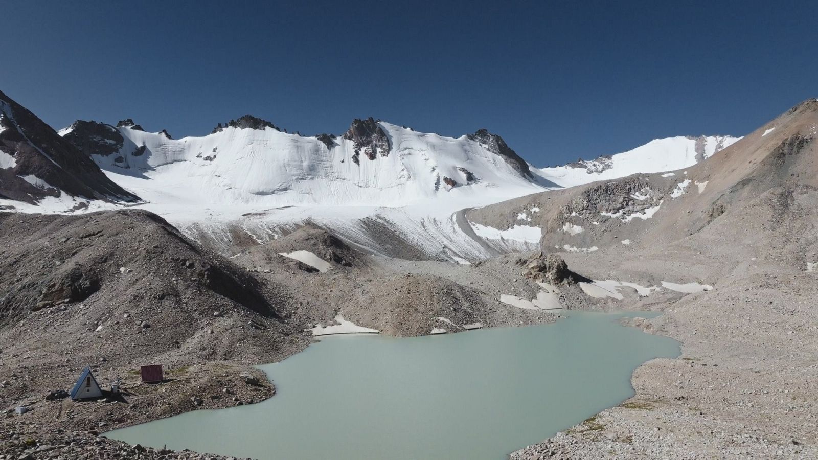 Paisaje de montaña con lago glaciar turquesa, terreno rocoso, tienda de campaña azul y blanca, estructura roja y picos nevados bajo cielo azul.