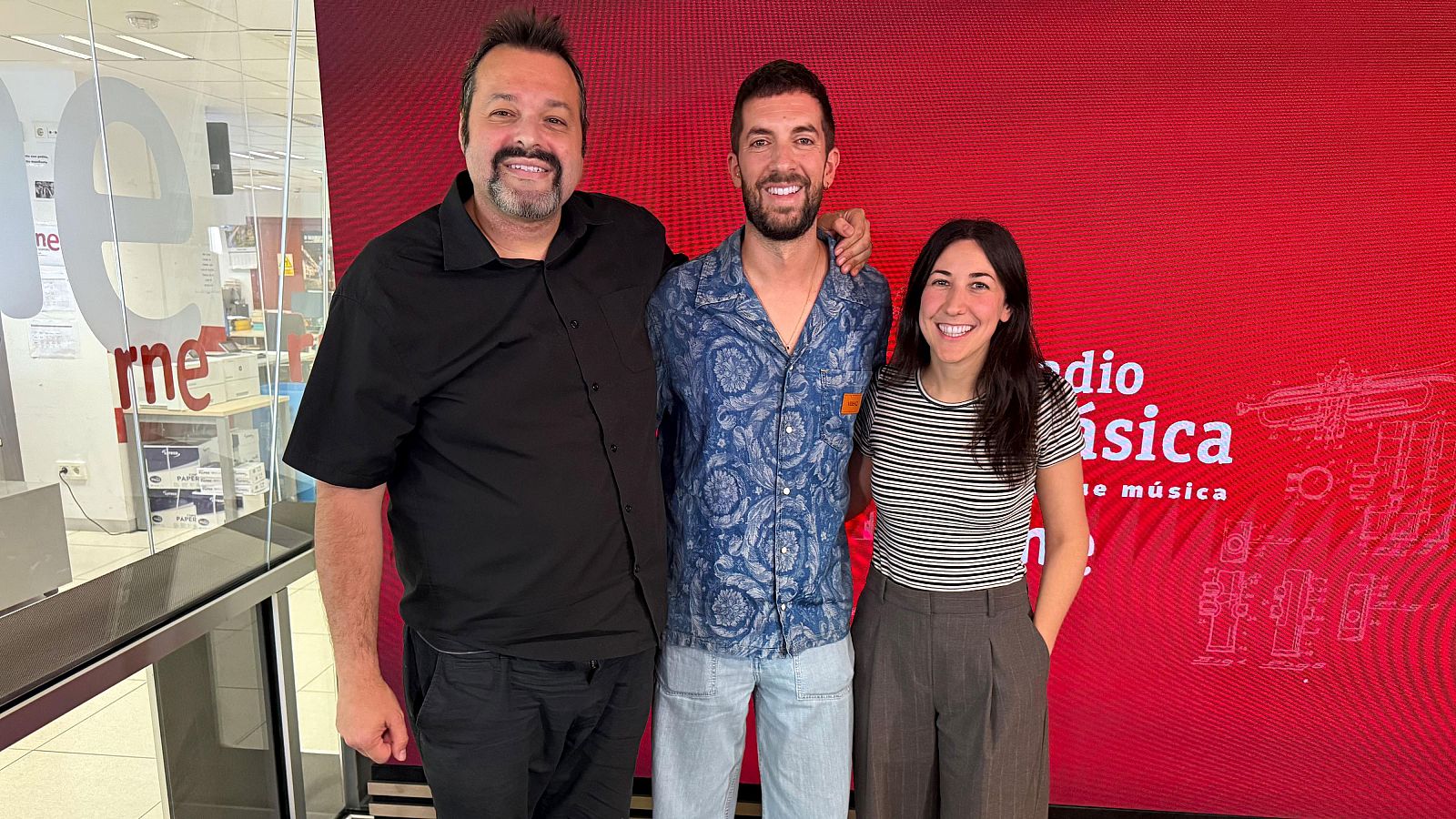Martín Llade, David Broncano y Clara Corrales sonríen en un estudio de Radio Clásica con fondo rojo y dibujos de instrumentos musicales. Se aprecia parte del logo de "rne" en un cristal.