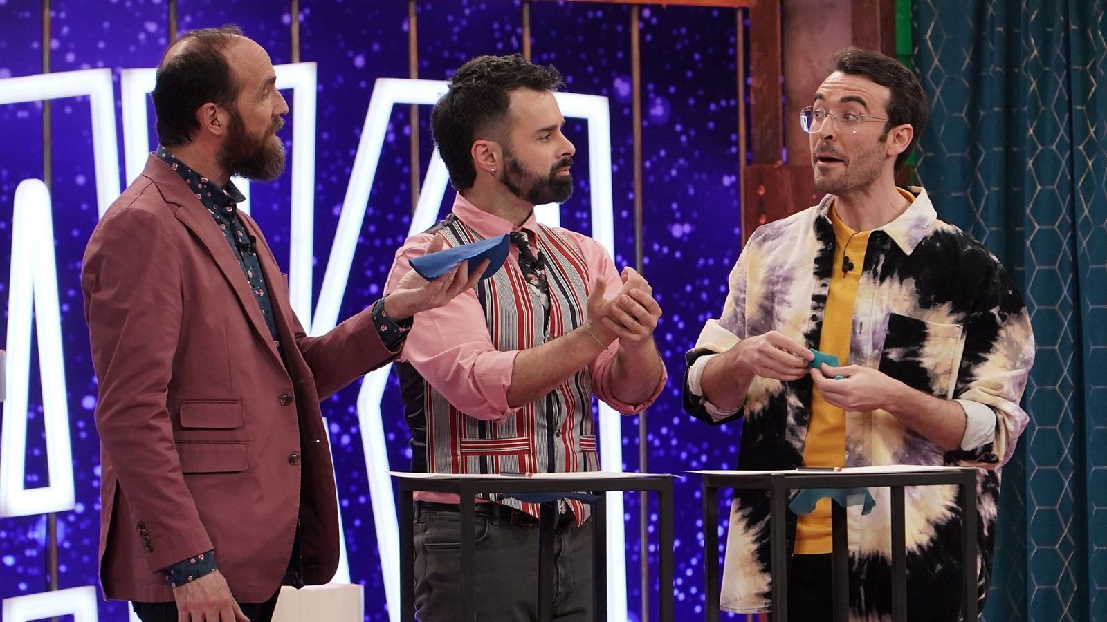 Ángel Martín, Goyo Jiménez y Javier Cansado en un plató con fondo azul y luces de neón. Goyo y Javier sostienen objetos azules.